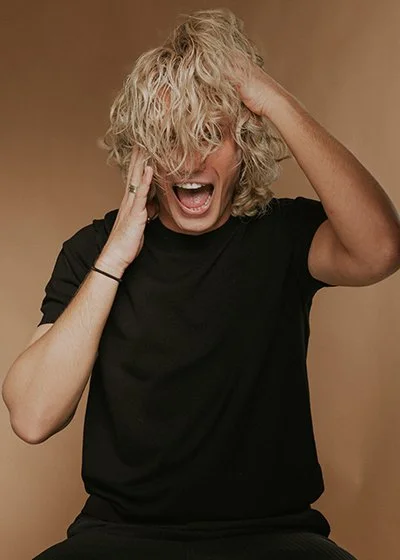 Ein Mann mit lockigem, blondem Haar, die offensichtlich freudig überrascht ist, die Hände am Kopf und den Mund weit geöffnet. Nico Kallisch Master Stylist und Co-CEO im HairClub Bad Oeynhausen, Model und Starfriseur, Promifriseur. Bester Friseur.