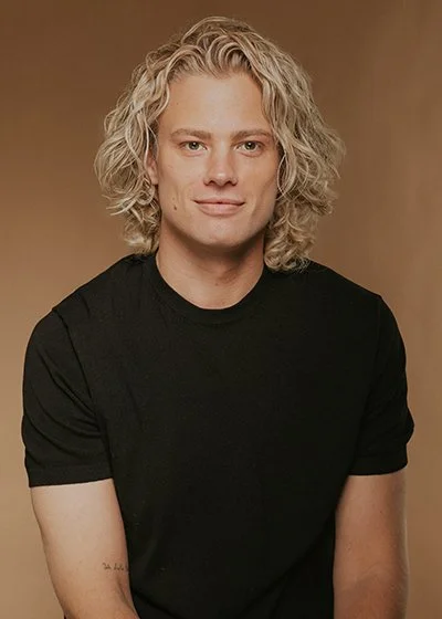 Porträt eines jungen Mannes mit langen, lockigen, blonden Haaren, schwarzen T-Shirt, vor braunem Hintergrund. Nico Kallisch Master Stylist und Co-CEO im HairClub Bad Oeynhausen, Model und Starfriseur, Promifriseur. Bester Friseur.