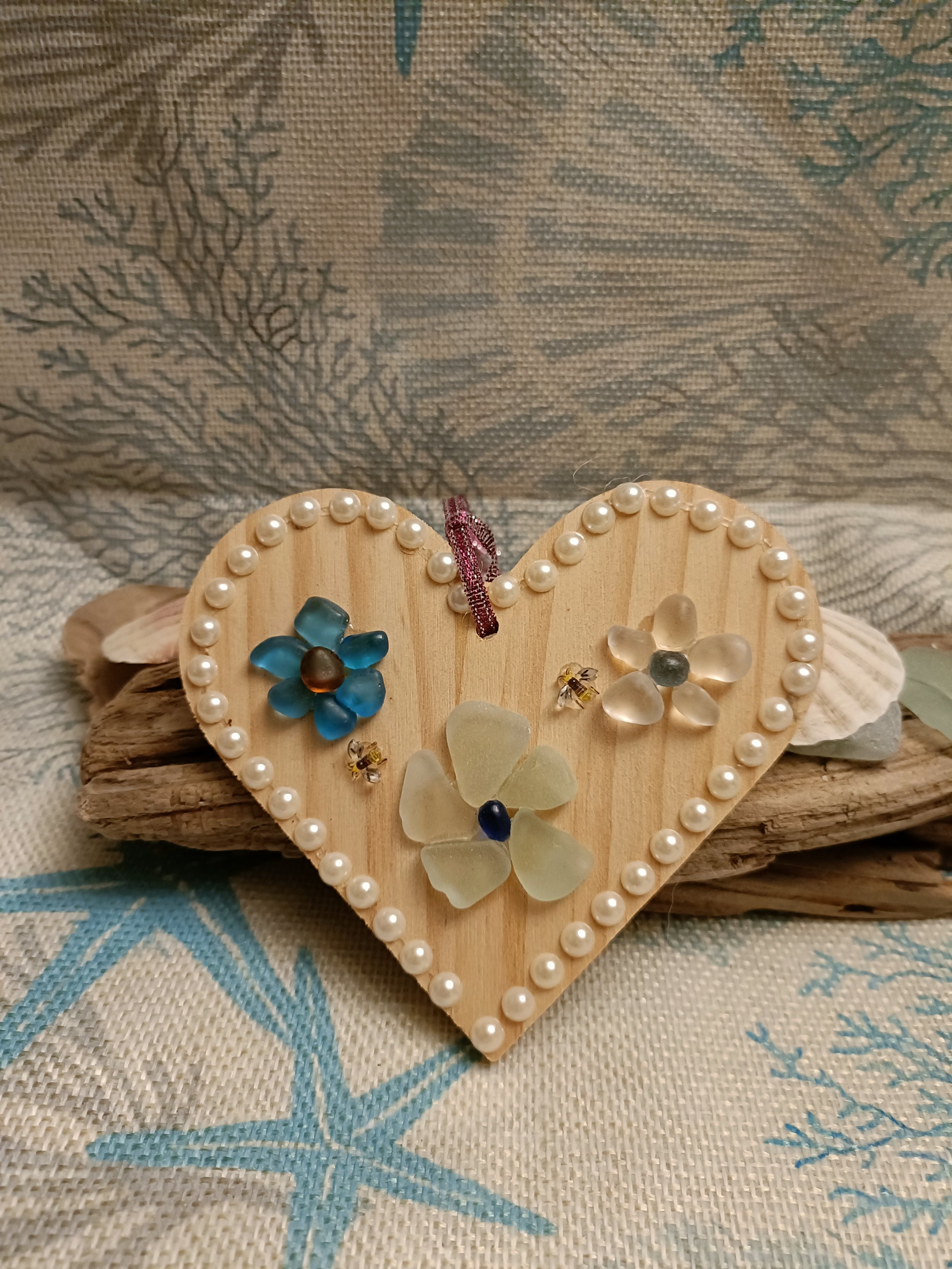 Heart-shaped wooden decoration with pearls and sea glass flowers, placed on a piece of driftwood against a marine-themed fabric background.