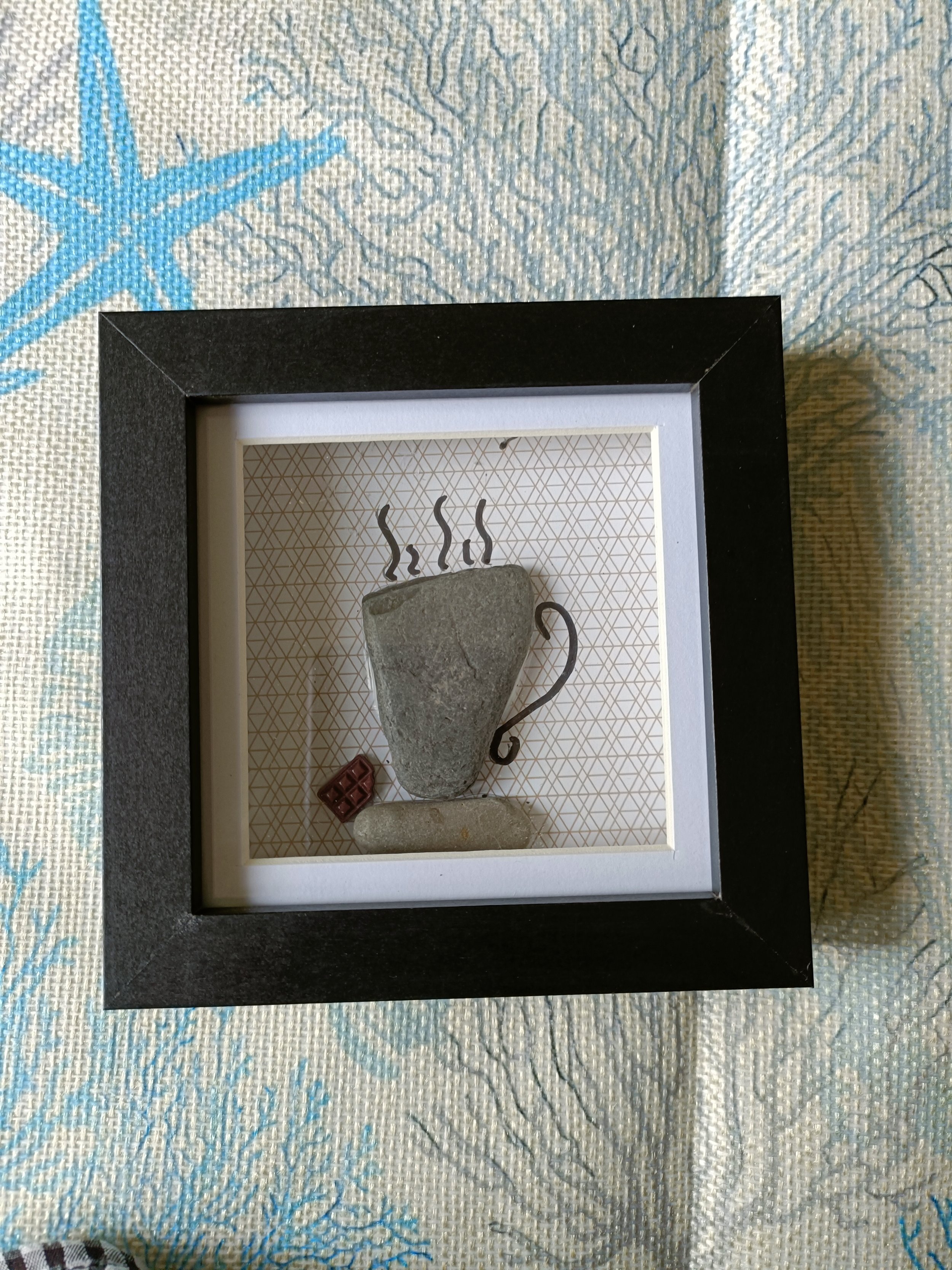 Framed artwork depicting a stone coffee cup with steam lines above, a small chocolate piece, and a biscuit on textured background, set on a patterned tablecloth.