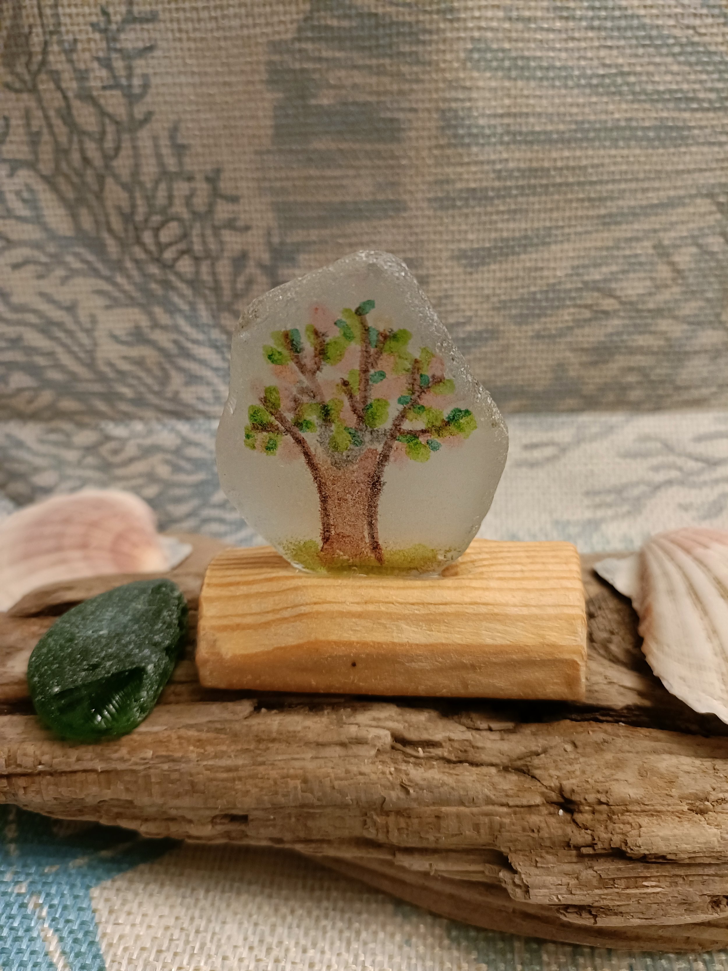 Small painted tree on glass, displayed on wooden block with seashells and driftwood in background.