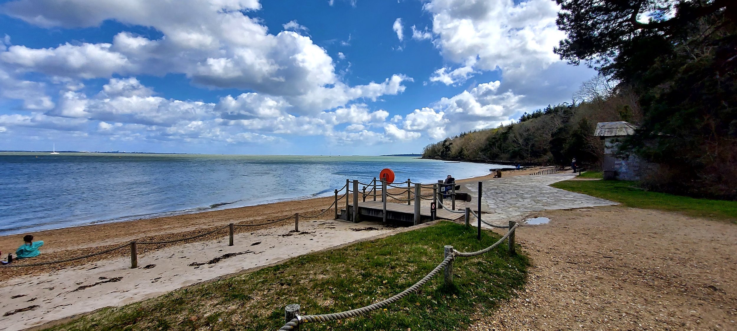 Beach with blue sky, people relaxing, trees, and a lifebuoy on a wooden platform.