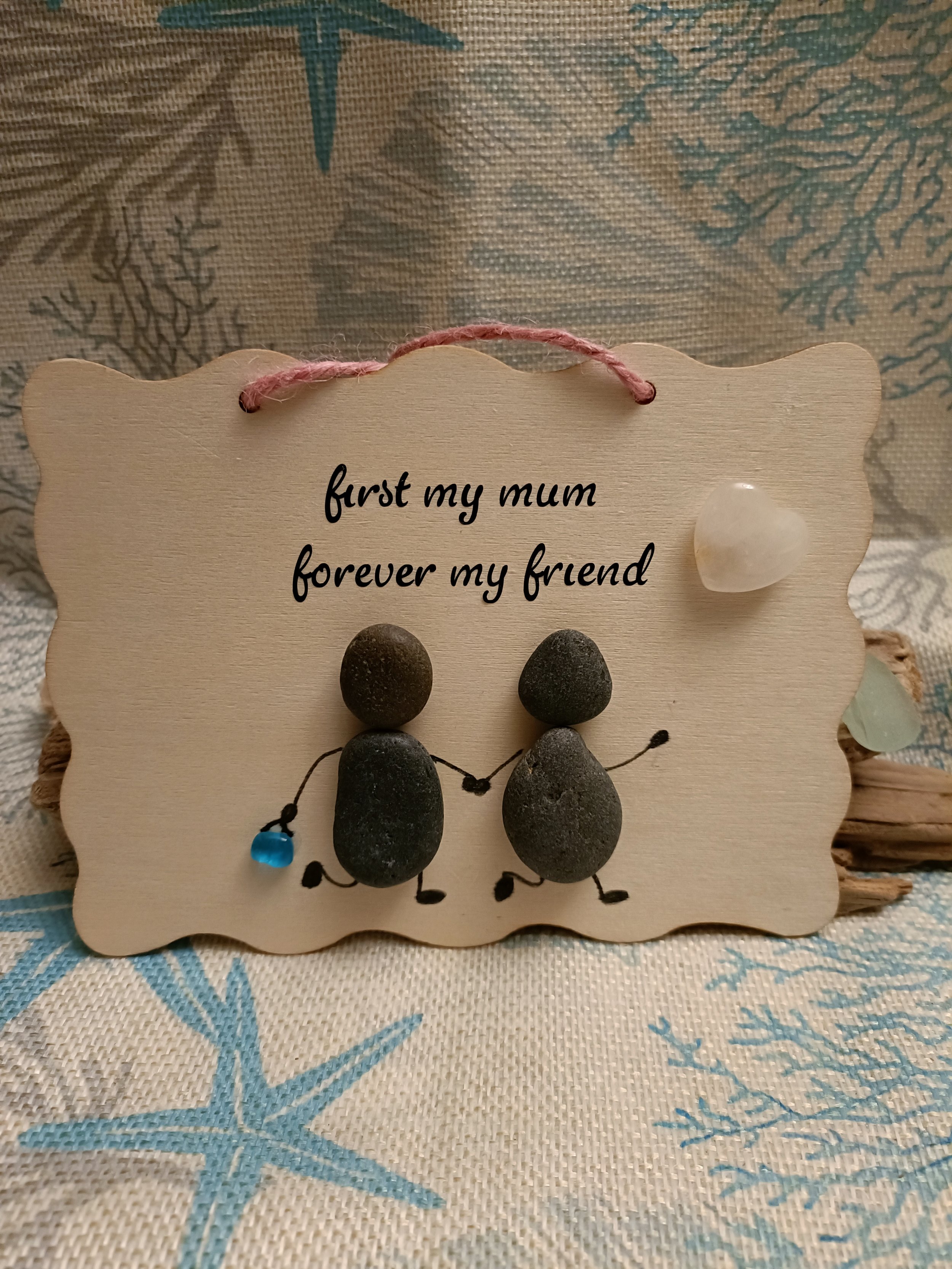 Decorative wooden plaque with pebbles forming two figures holding hands. Text reads "first my mum forever my friend." A small heart-shaped decoration is attached.
