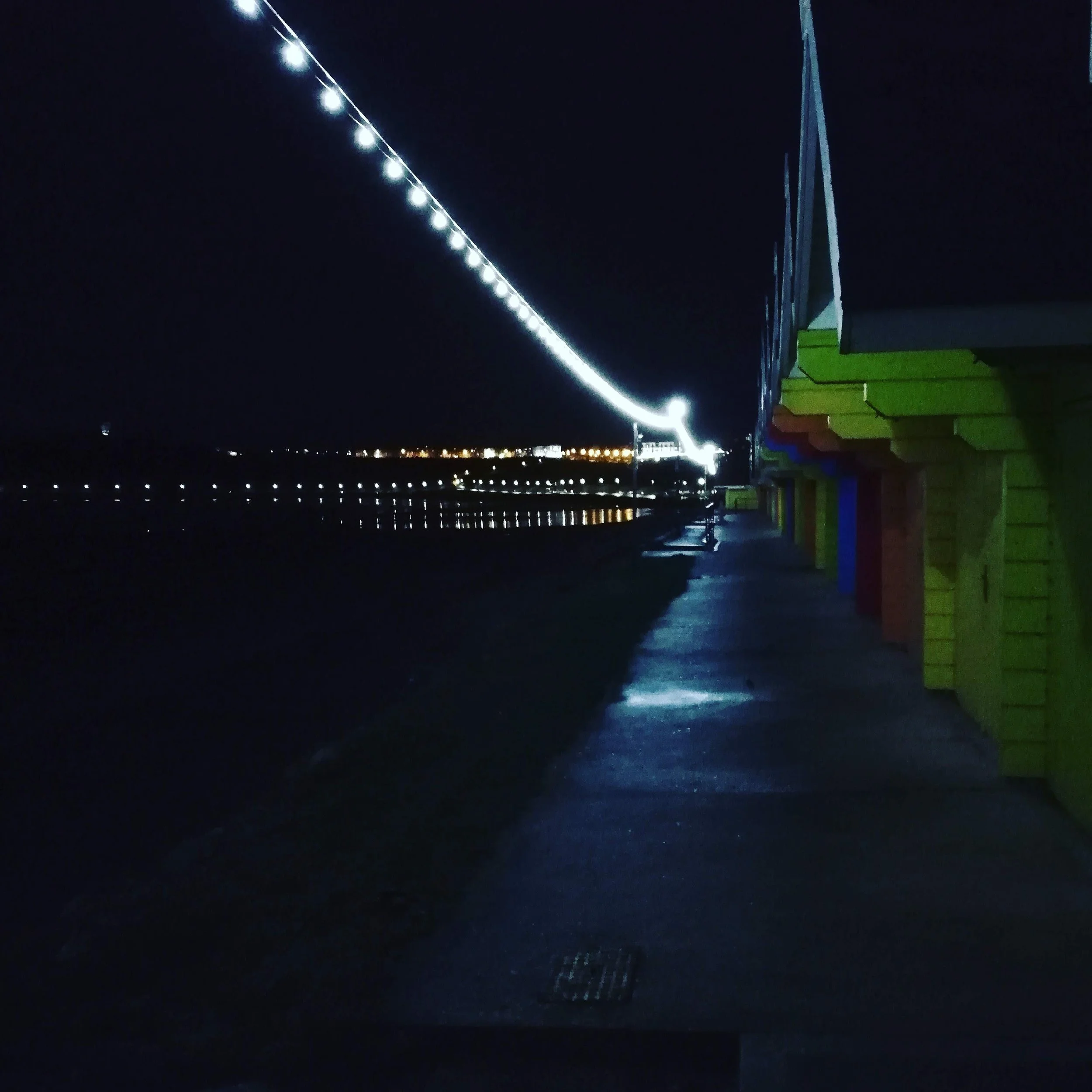 Night scene with colorful beach huts and string lights along a promenade, possible water reflections.