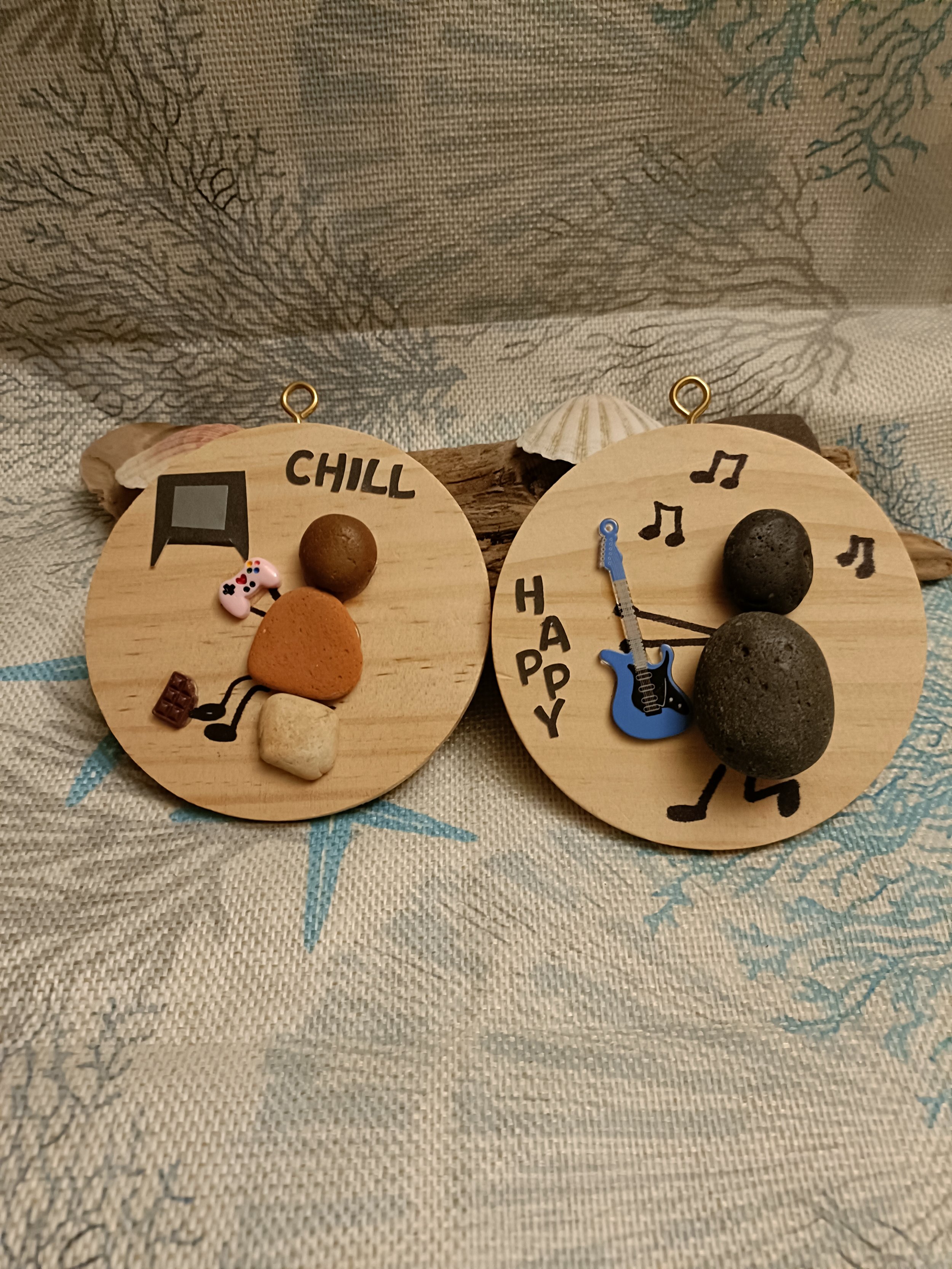 Two round wooden ornaments with small rocks and decorations; one with 'CHILL' and a TV and game controller, the other with 'HAPPY,' musical notes, and a guitar; backgrounds feature shells and a patterned cloth.