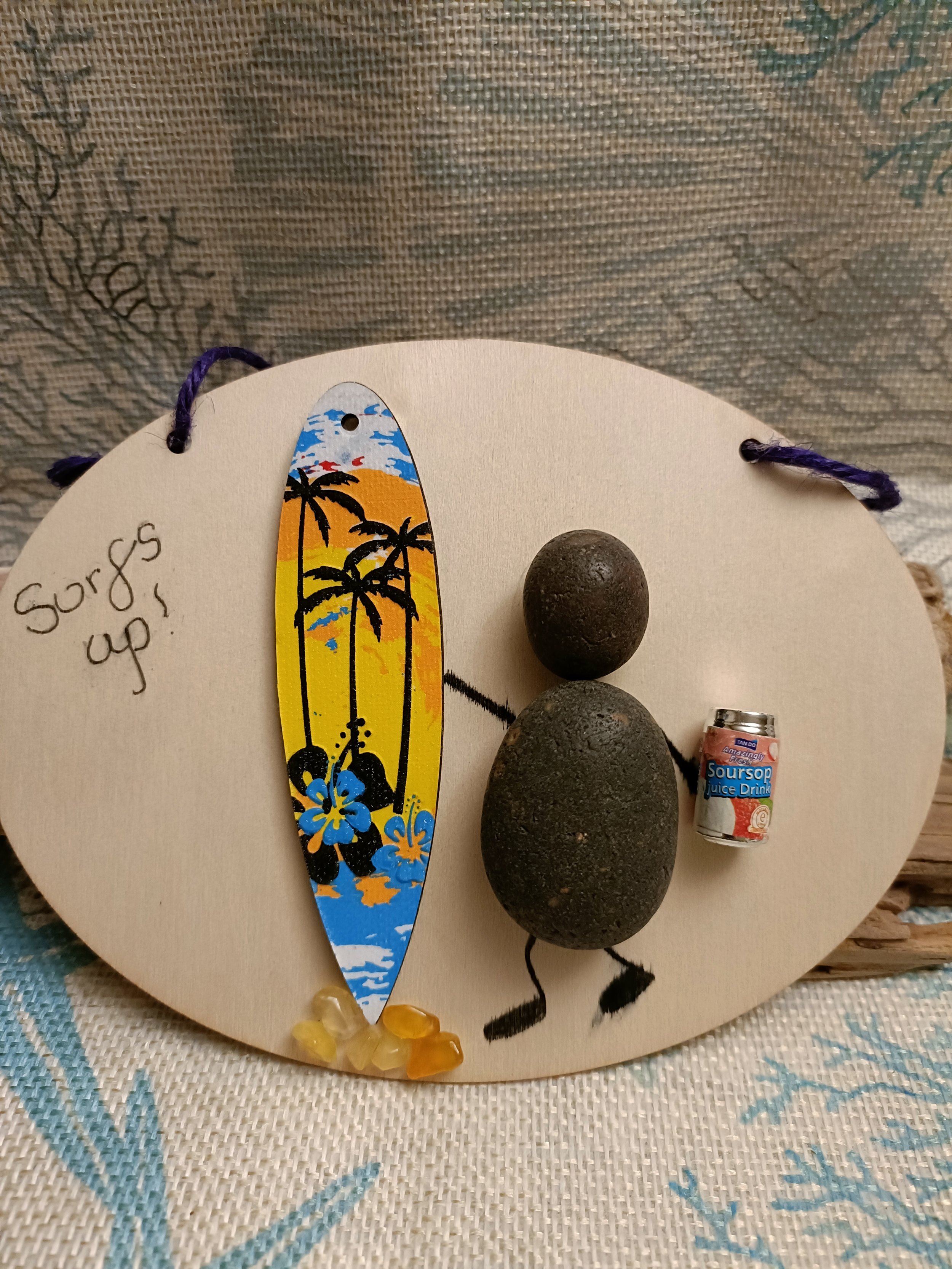 A decorative surf-themed ornament featuring a figure made from stones, holding a small can labeled "Soursop Juice Drink," alongside a colorful, miniature surfboard with a palm tree and sunset design. The text "Surf's up!" is written on the wooden bas