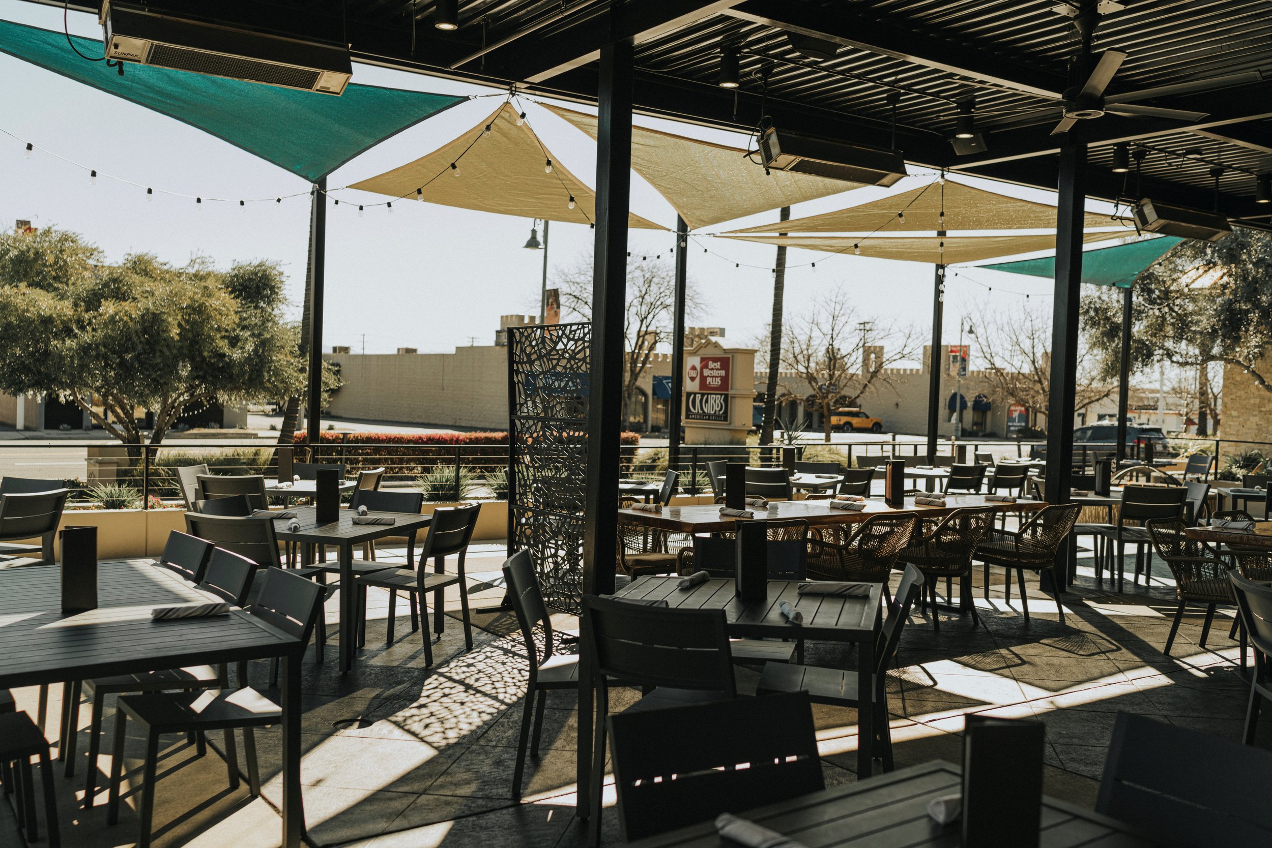Outdoor restaurant patio with tables and chairs, shaded by triangular canopy sails, with trees and a street view in the background.