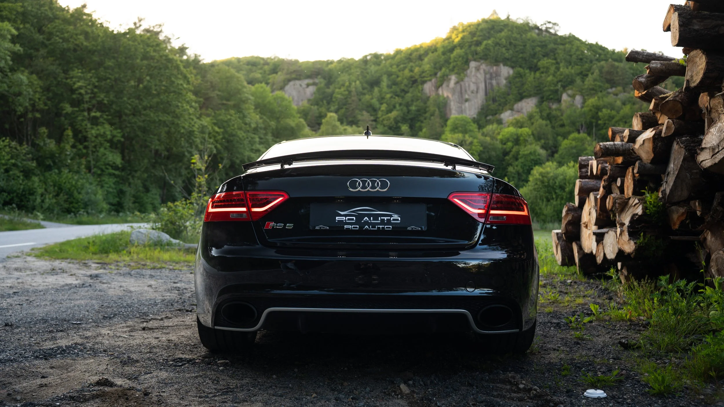 Sort sportbil parkert på en grusvei med grønne trær og fjell i bakgrunnen.