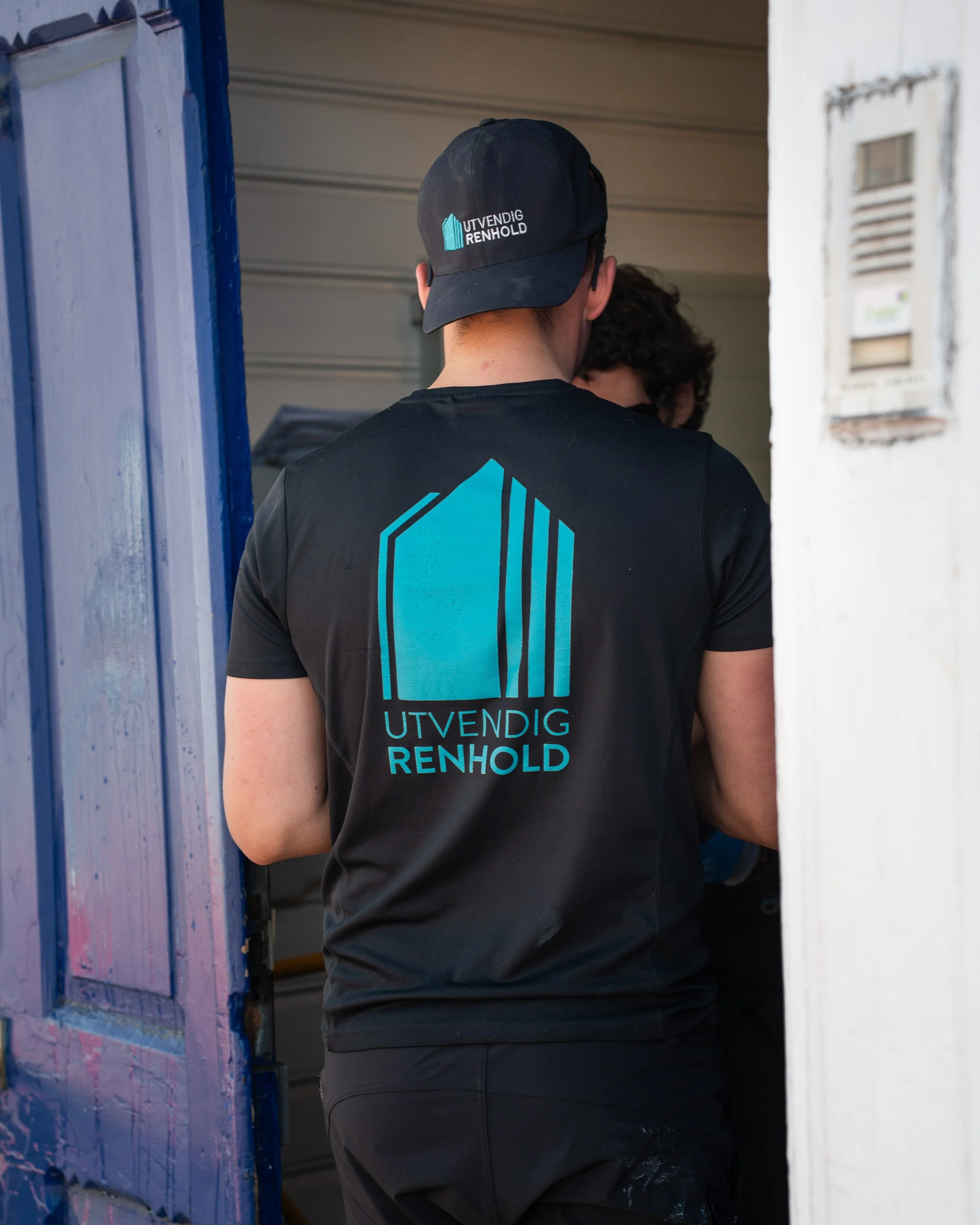 En person i svart uniform med logo og tekst 'UTVENDIG RENHOLD' på ryggen, står i en inngang, antydet til å utføre rengjøring.