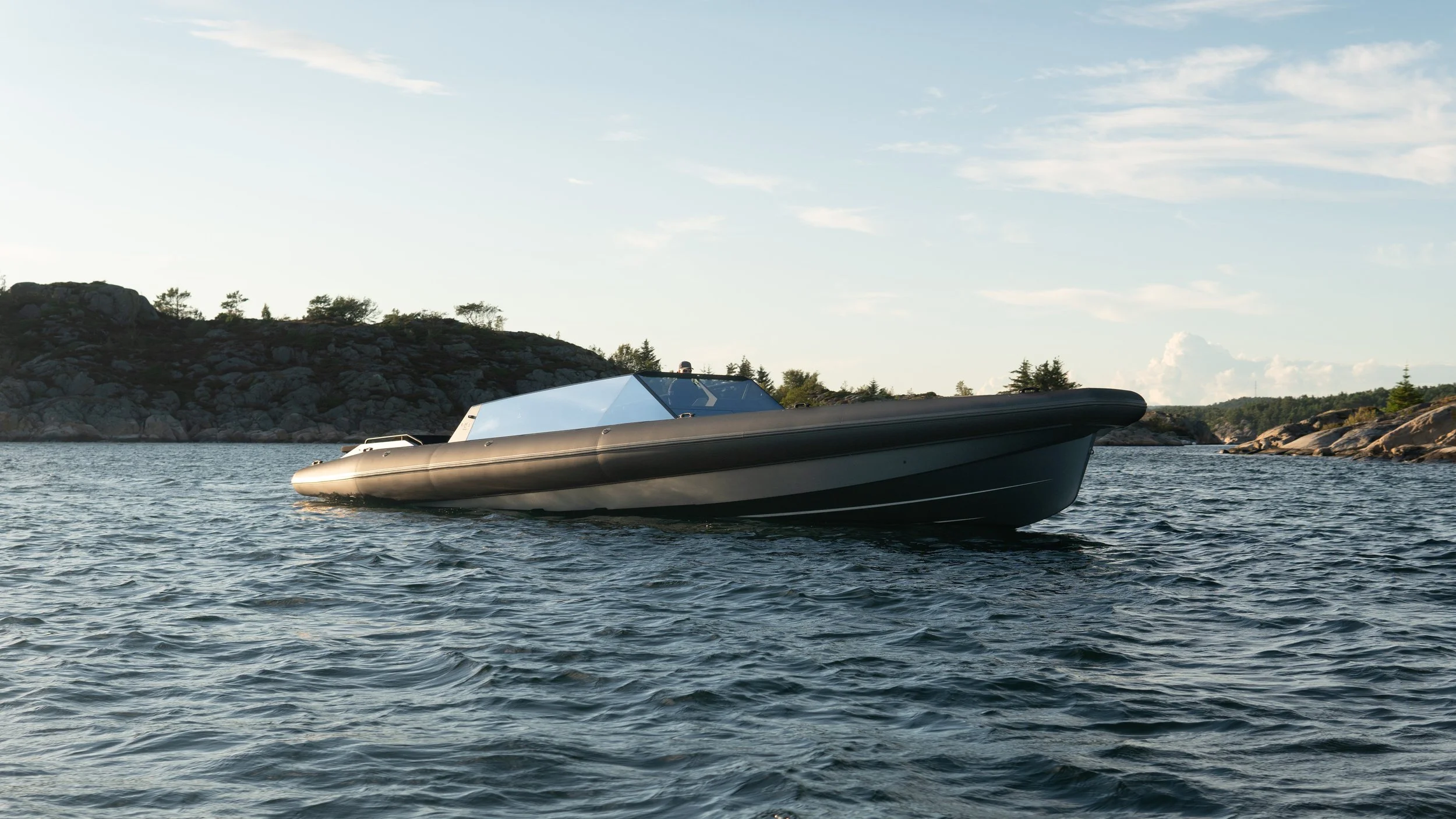 En moderne motorbåt på vannet med skjær og trær i bakgrunnen.
