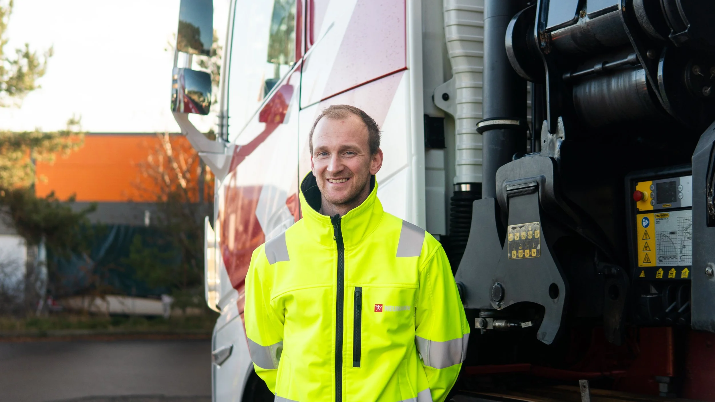 En mann i en høyt synlig gul vest står foran en stor brannbil, smilende mot kameraet. Bak ham er noe trær og en bygning med oransje vegg, utendørs.