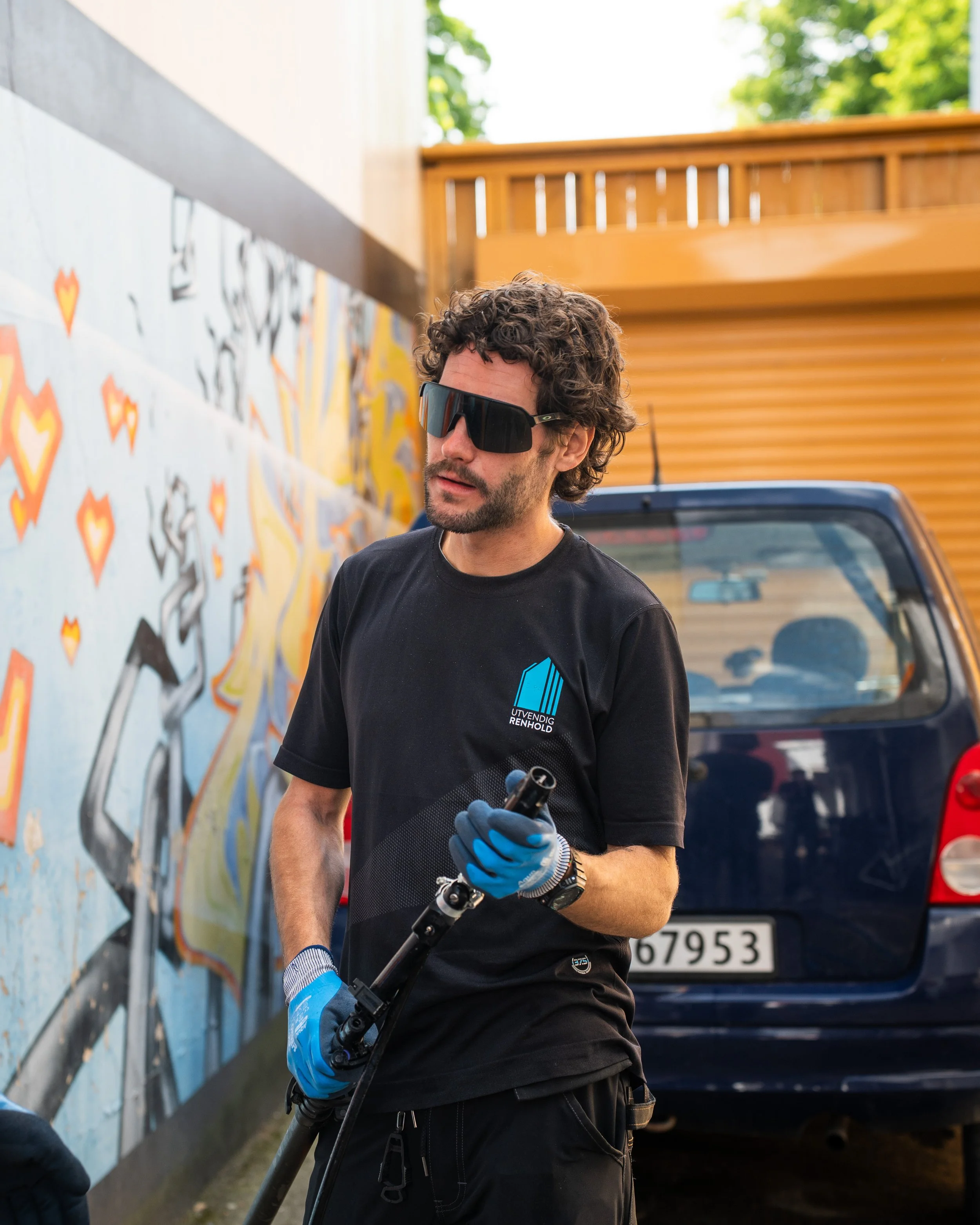 En mann med krøllete hår og solbriller holder en høytrykksspyler, står foran en vegg med graffiti og en blå bil.