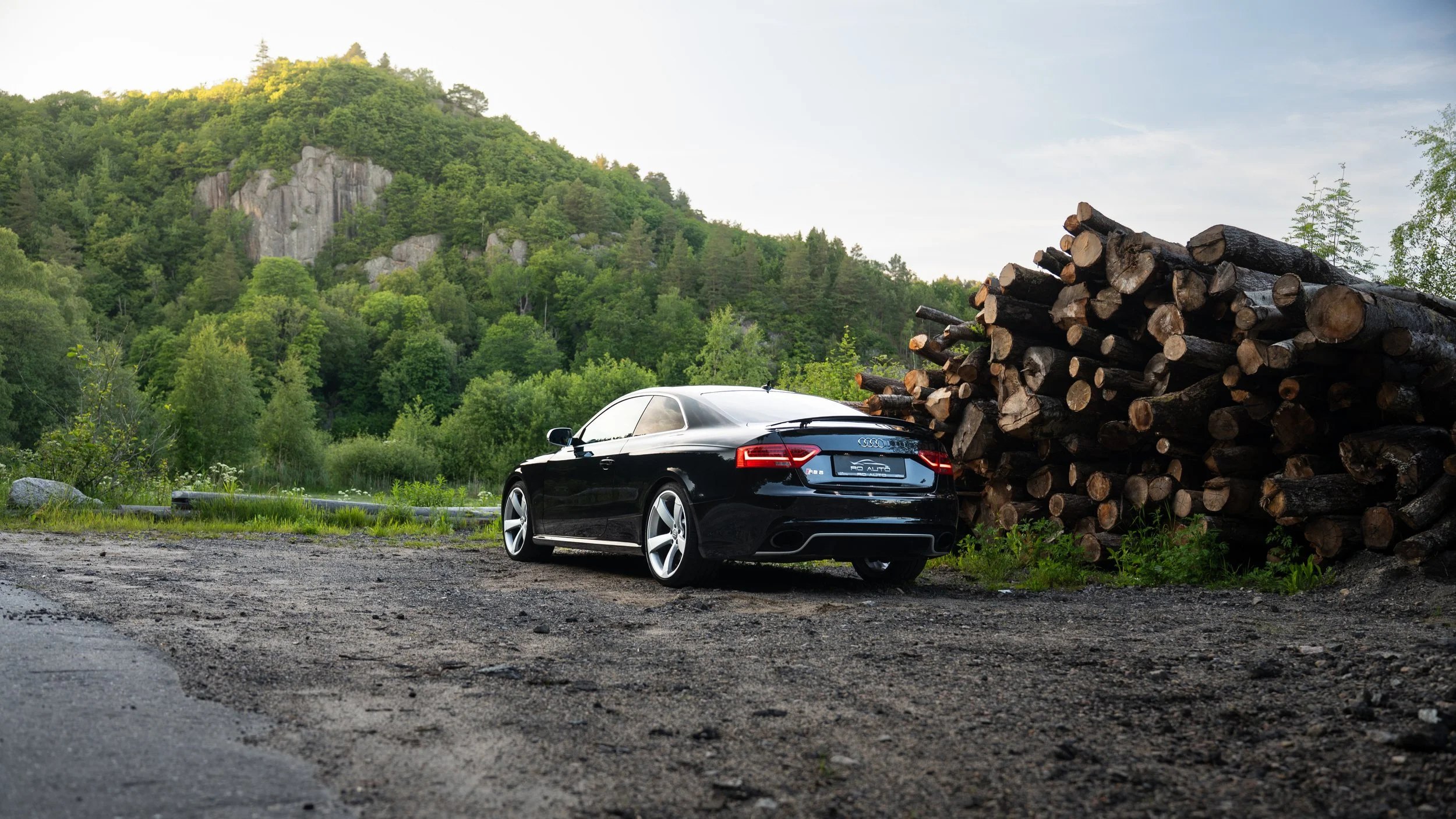 En svart Audi-sportbil parkert ved siden av en stor stab av vedstokker i en skogkledd fjellområde.