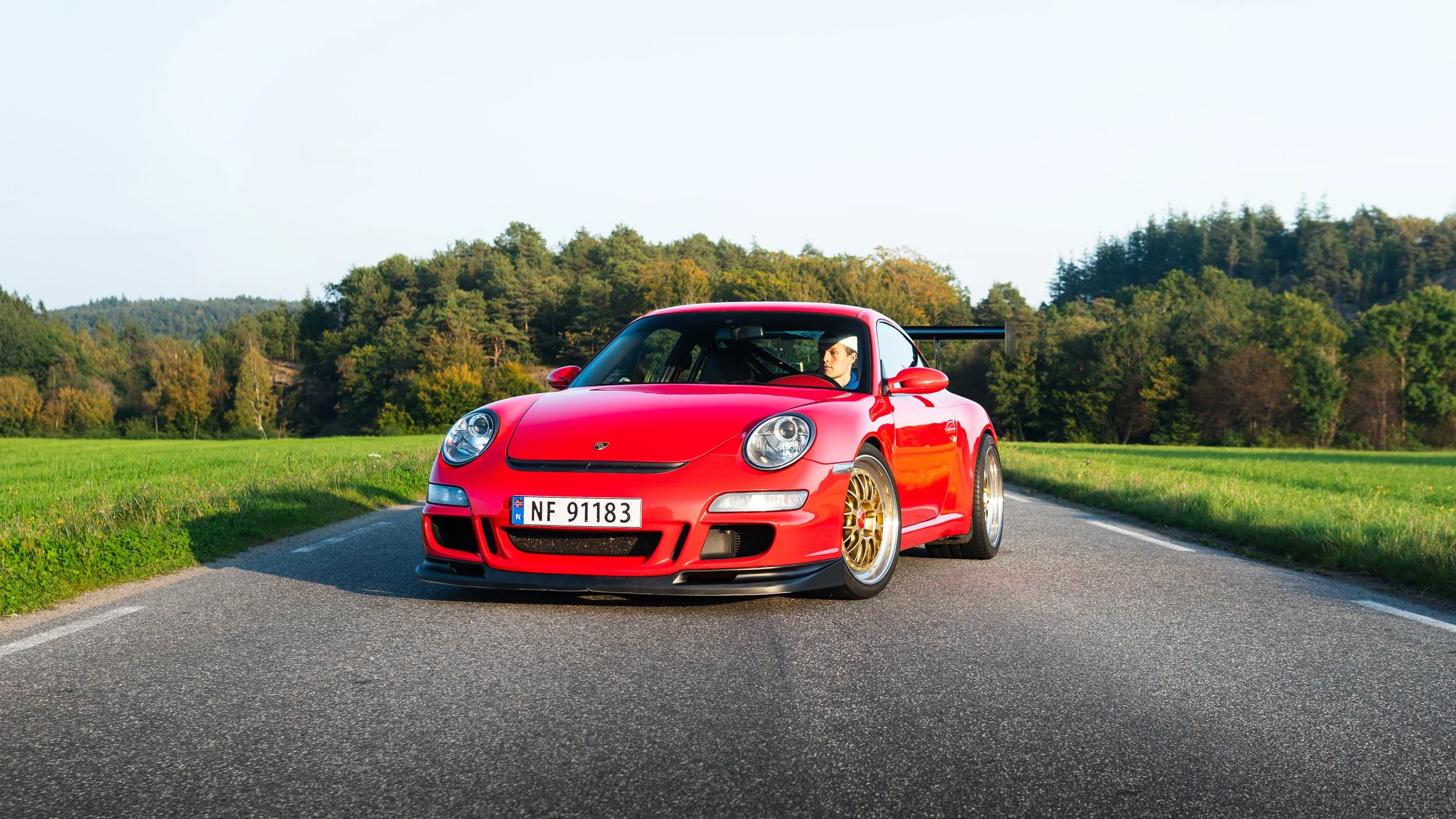 Rød sportsbil på en landevei med grønt landskap og trær i bakgrunnen.