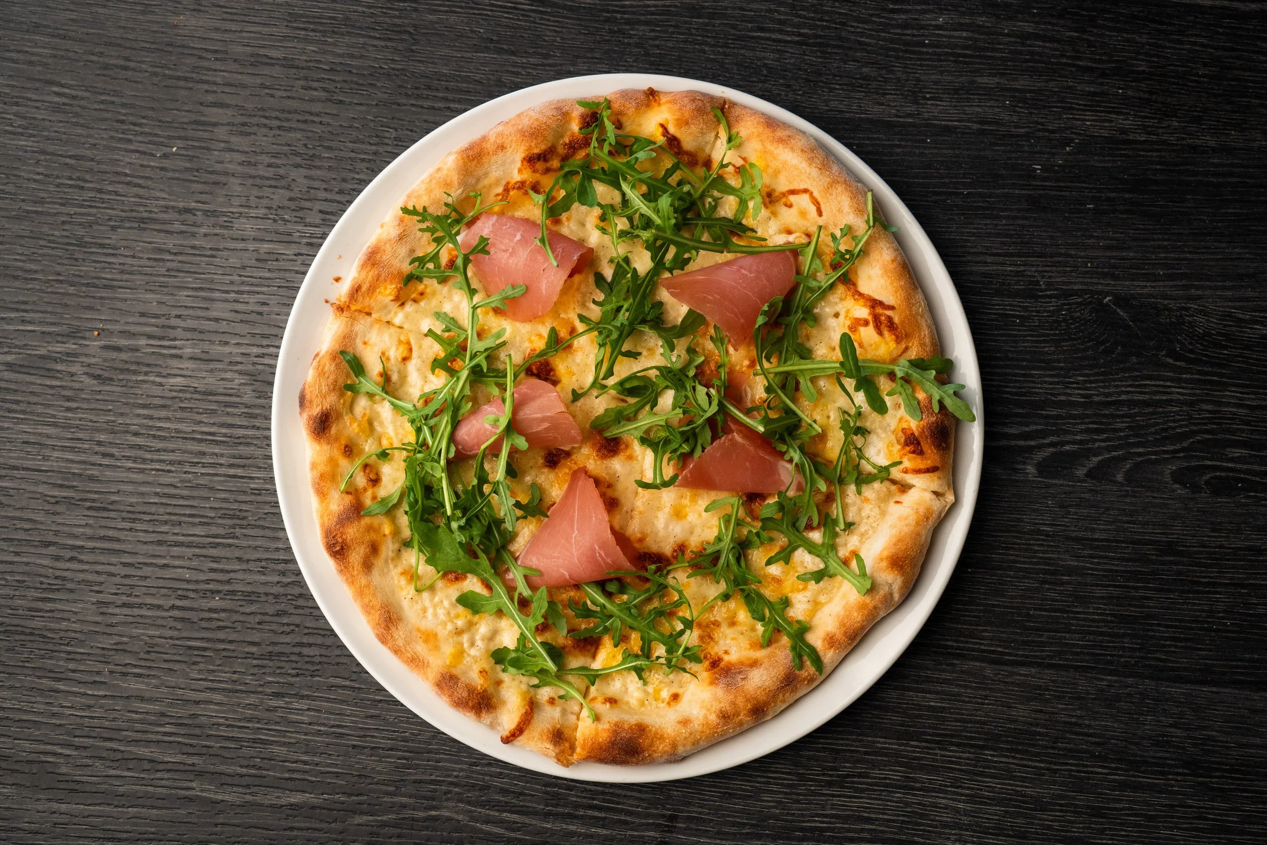 Pizza med ost, kokt skinke og ruccola på en svart trebord.