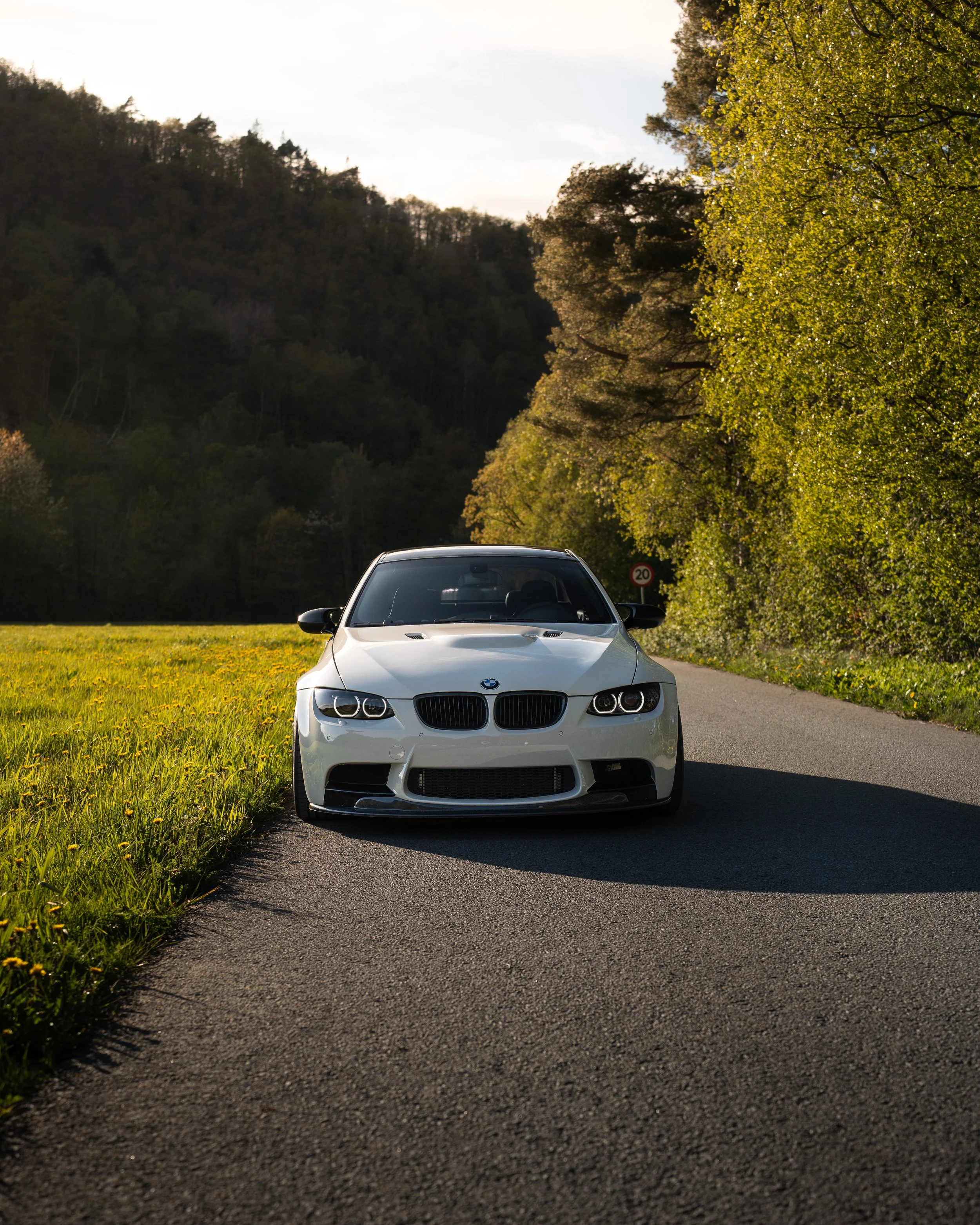 En hvit BMW kjører på en smal vei omgitt av grønne trær og åker med åpent landskap og fjell i bakgrunnen.