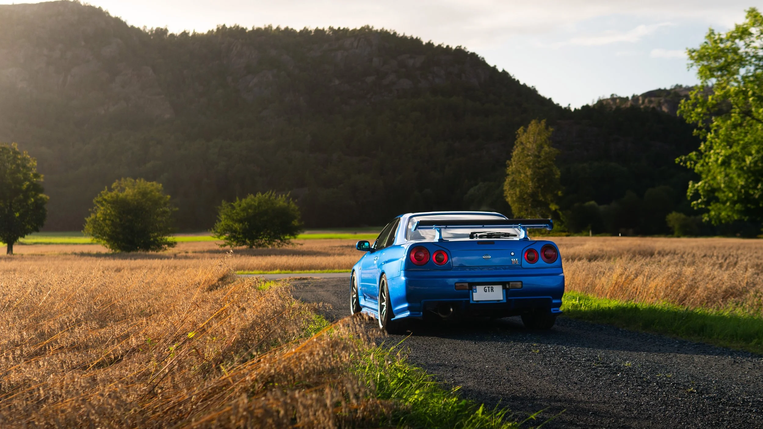 Blå sportsbil på en landevei omgitt av gress og trær, med fjell i bakgrunnen og solskinn.
