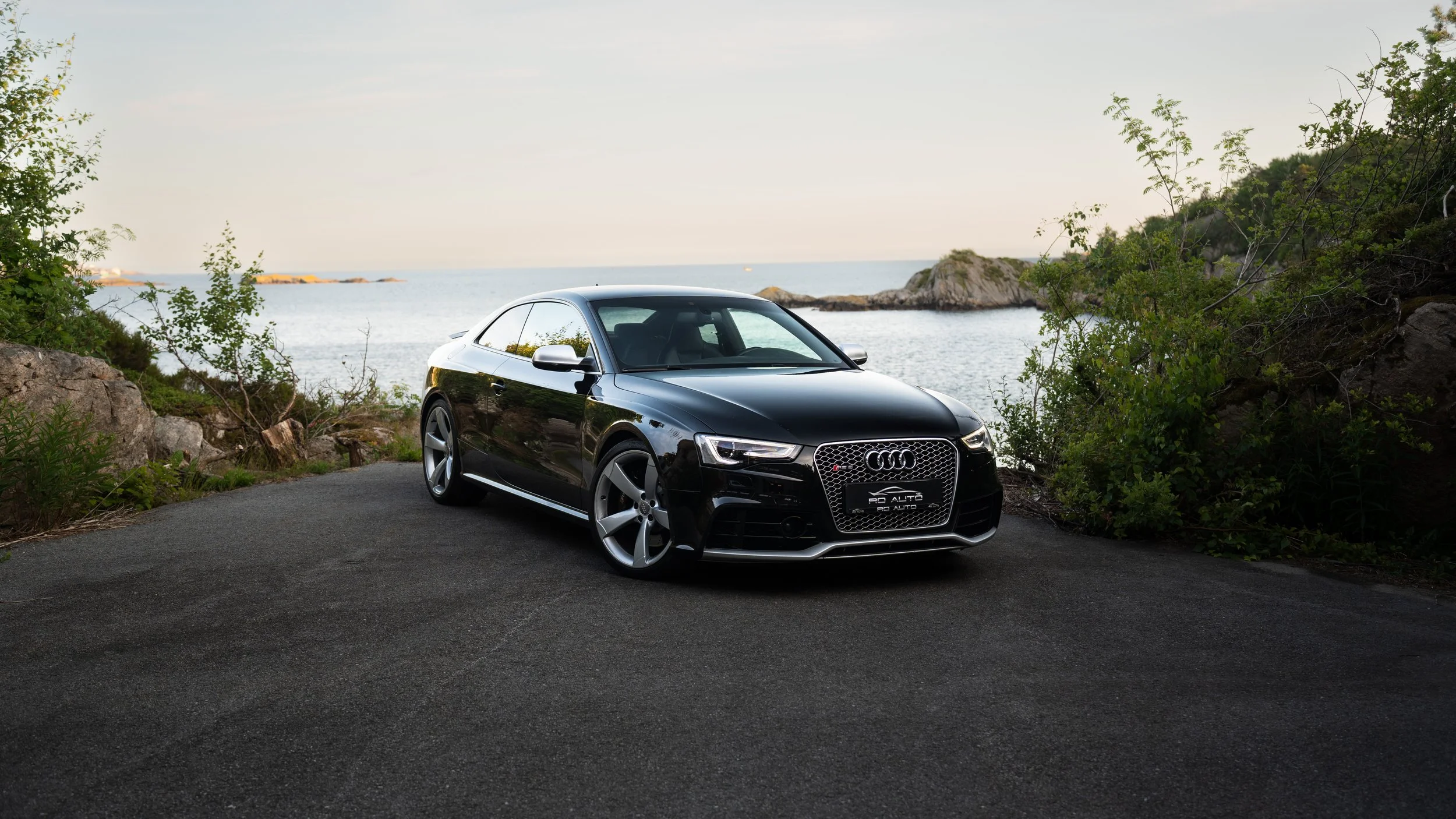 Sort Audi kupé parkert ved sjøen med steiner og grønt vegetation rundt og en fjord eller hav i bakgrunnen.