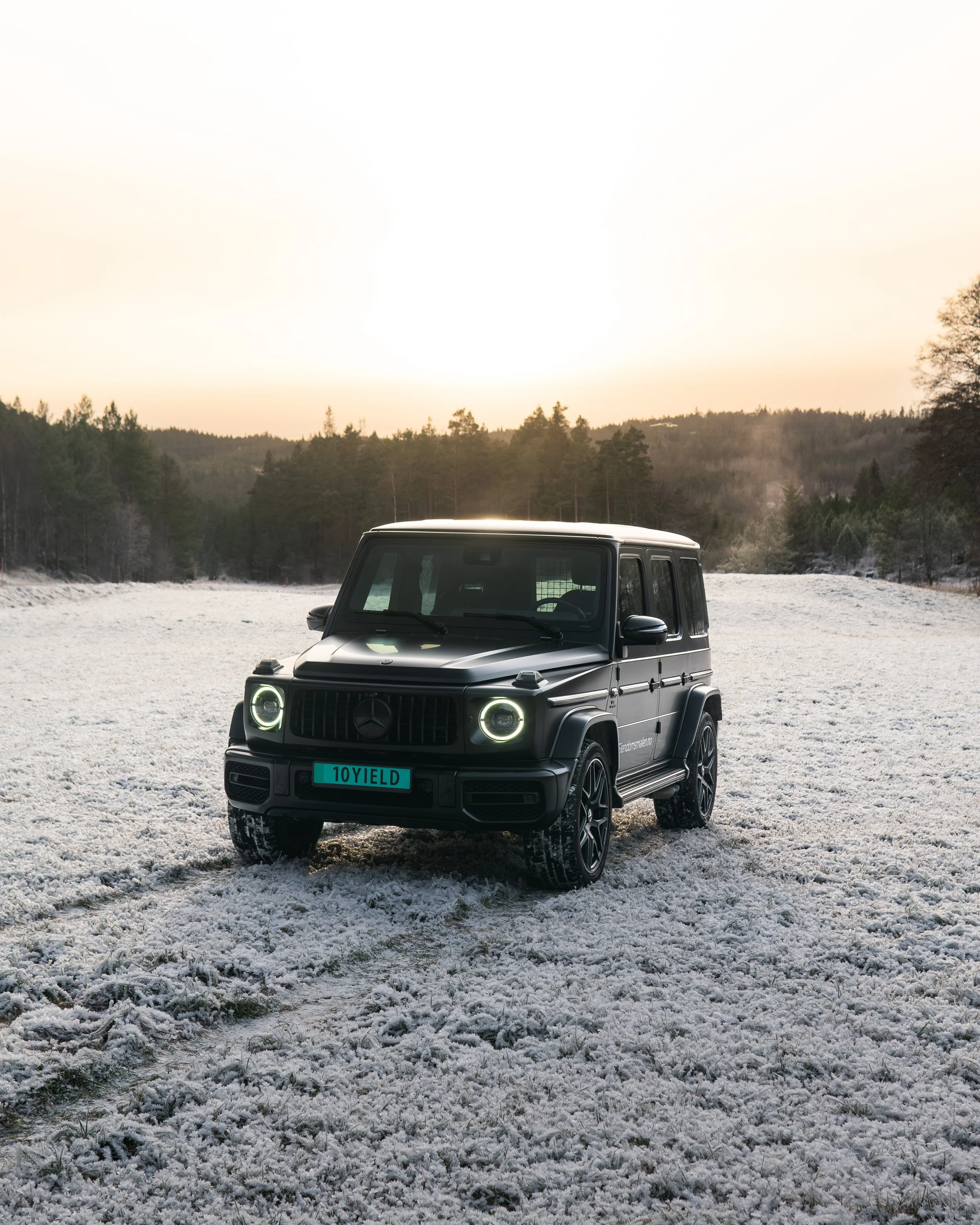 En svart Mercedes-Benz G-klasse SUV parkert på snødekt åpen slette med skog i bakgrunnen under en solnedgang.