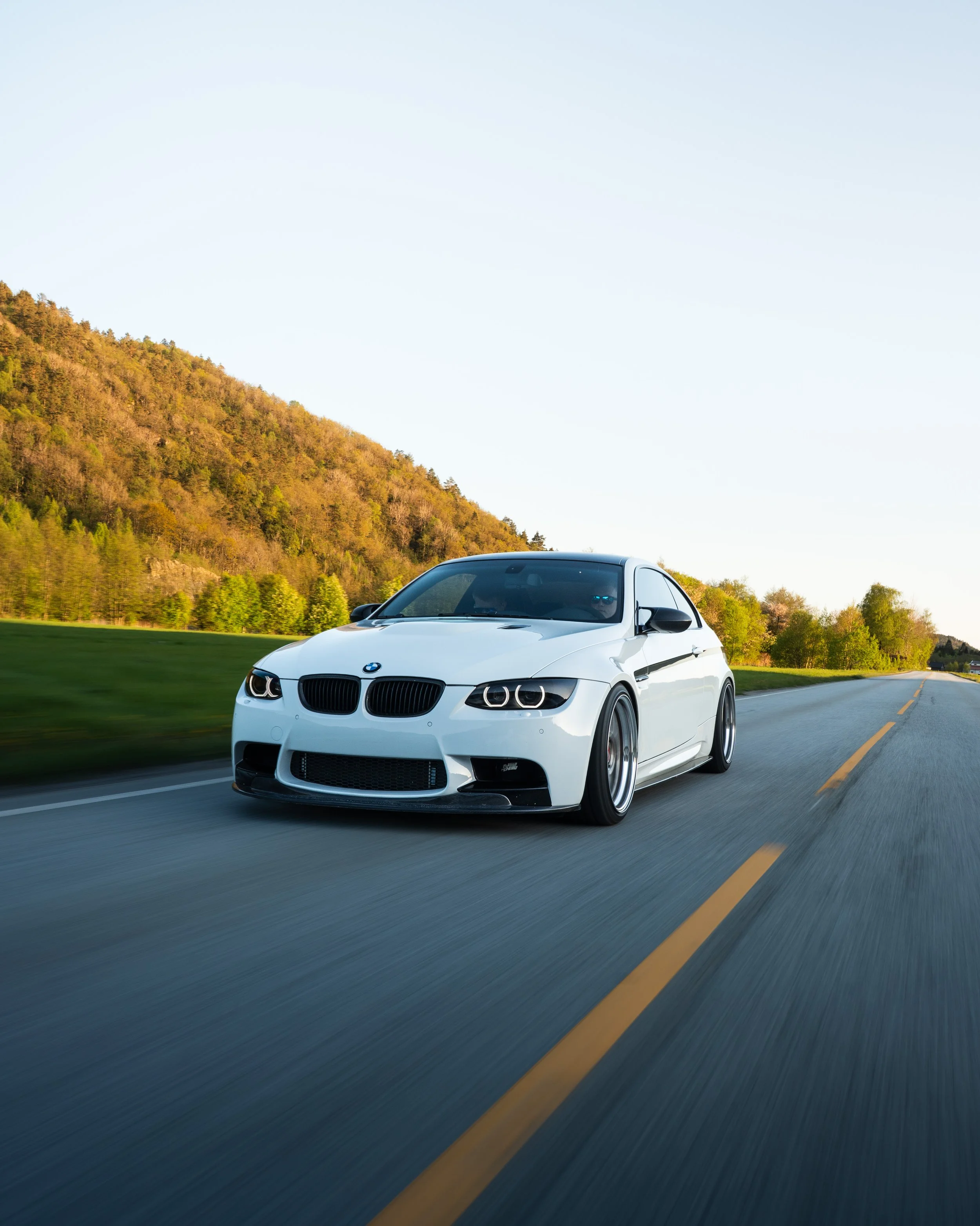En hvit BMW kjører på en tom landevei med grønne trær og en ås i bakgrunnen på kvelden.