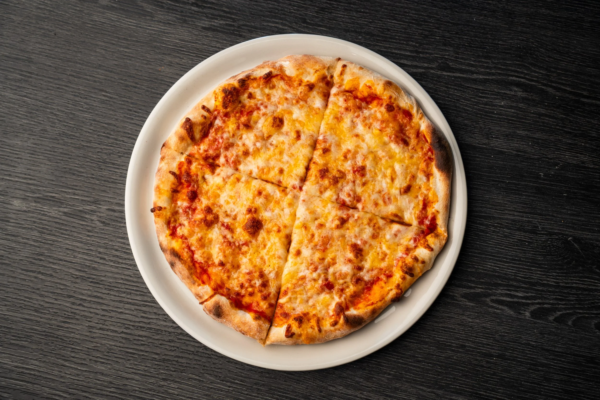 En pizza med ost og tomatsaus, skåret i fire stykker på en hvit tallerken, plassert på en svart trebord.