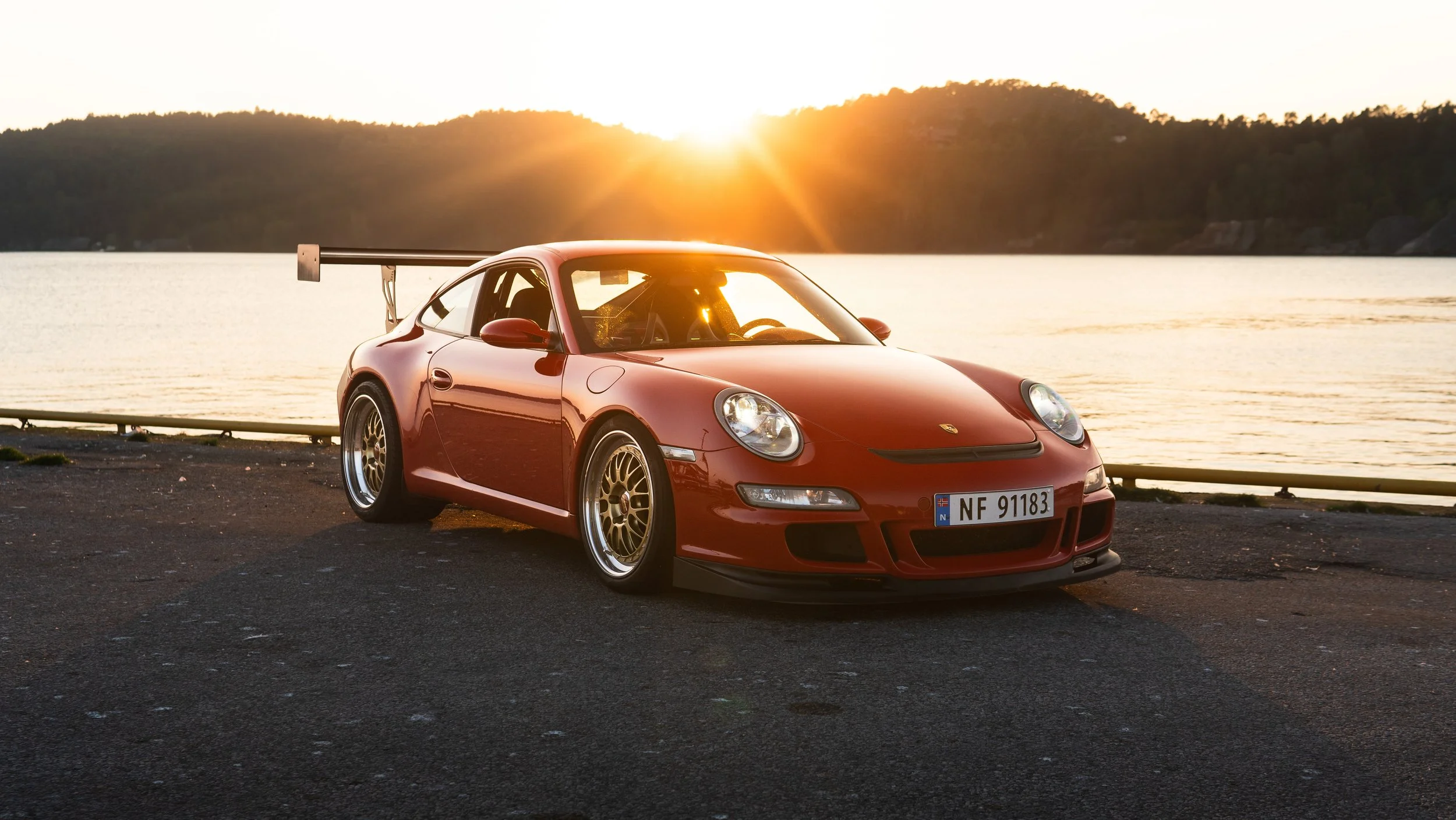 Rød Porsche sportsbil parkert ved vannet med fjell i bakgrunnen under solnedgang.