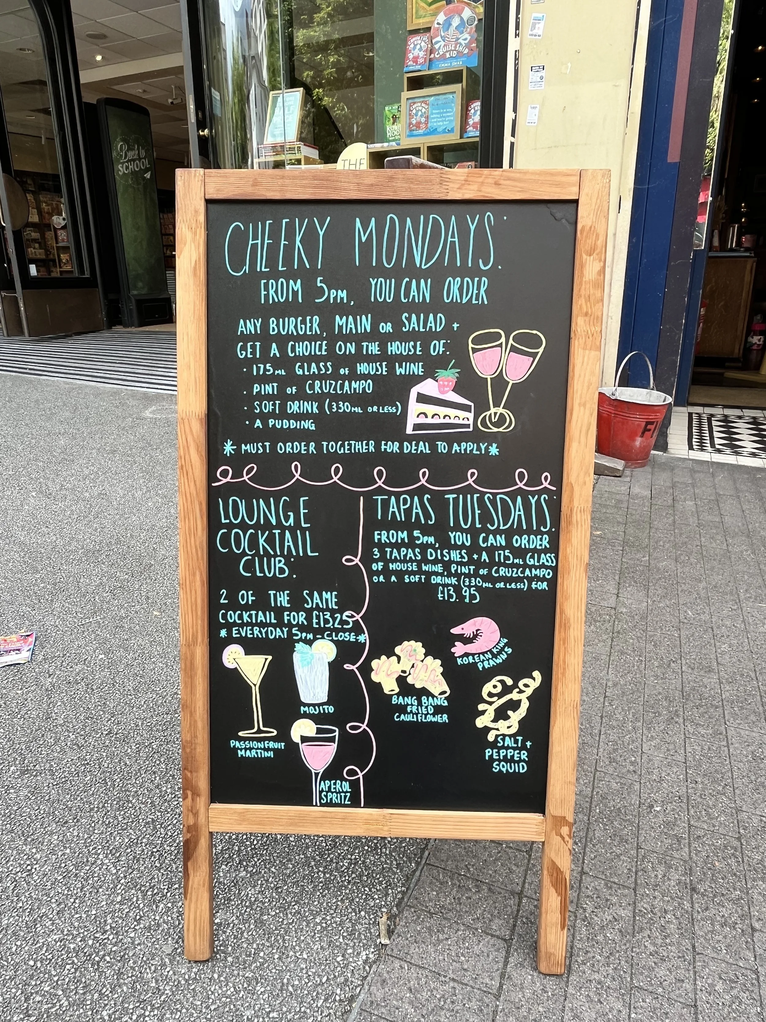 A board pavement sign promoting 'Cheeky Mondays', 'Tapas Tuesdays' and '2 for £13.25 cocktails after 5pm'. Hand Drawn text + image using Posca Markers (2025).