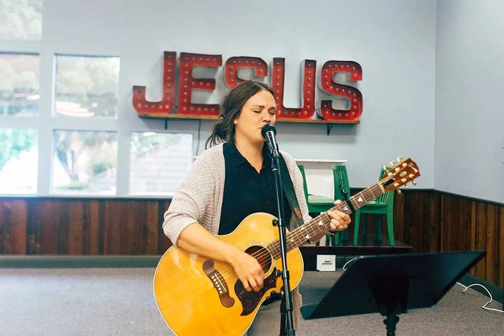 Woman singing and playing a guitar in front of a 'Jesus' sign