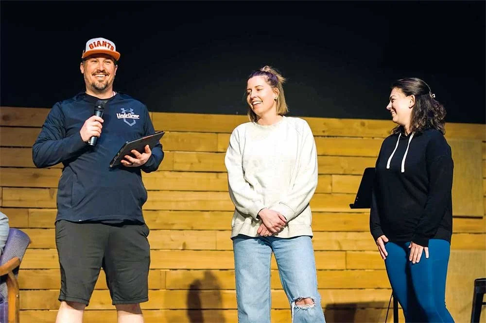 Man holding a mic and speaking on stage with 2 women laughing