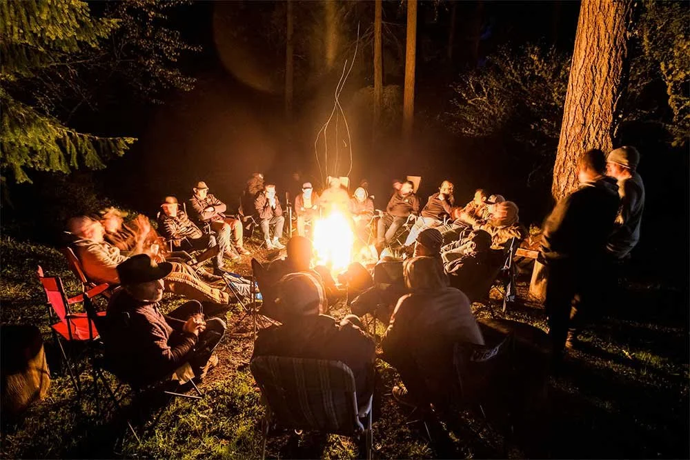 2 dozen men sitting in a circle around a campfire in the woods at night