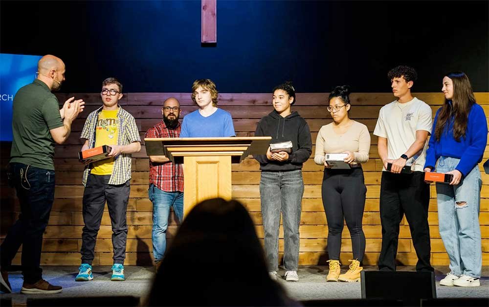 A group of seven young people standing on a stage receiving awards and recognition from an adult man.