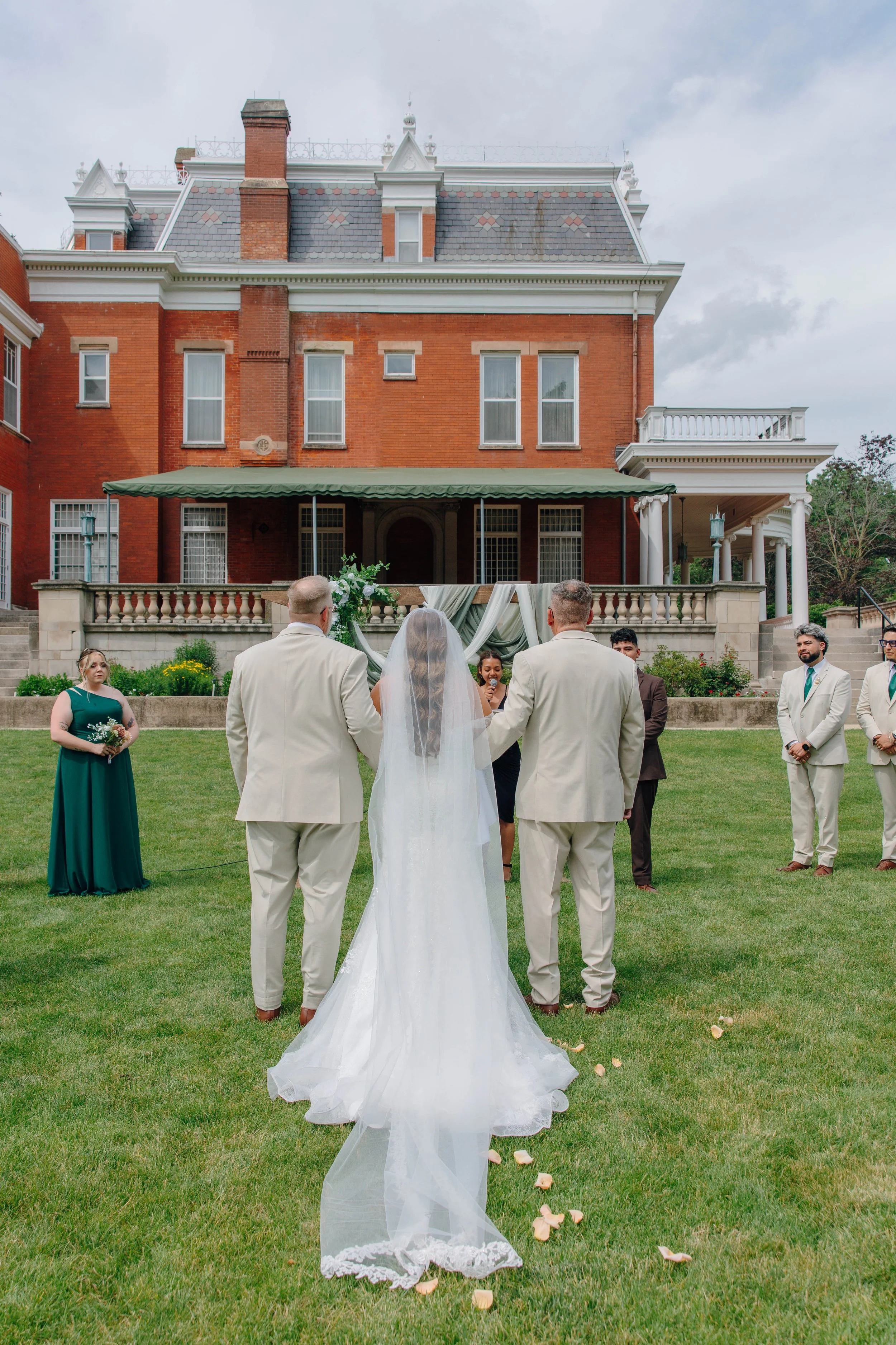 Wedding ceremony taking place outdoors on a lawn in front of a large brick mansion. The scene is decorated with flowers and fabric on a small platform, with rose petals scattered on the ground.