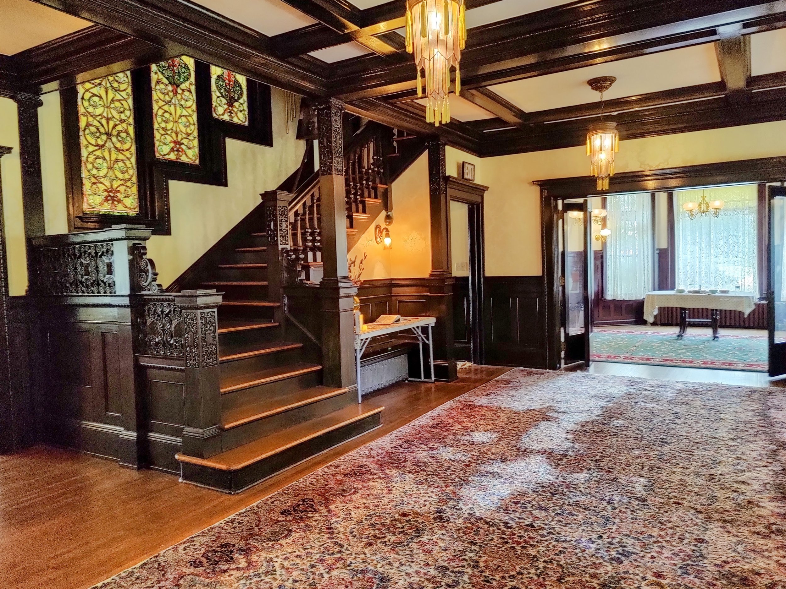 Luxurious vintage foyer with dark wood paneling, ornate staircase, stained glass windows, hanging chandeliers, and patterned rugs.