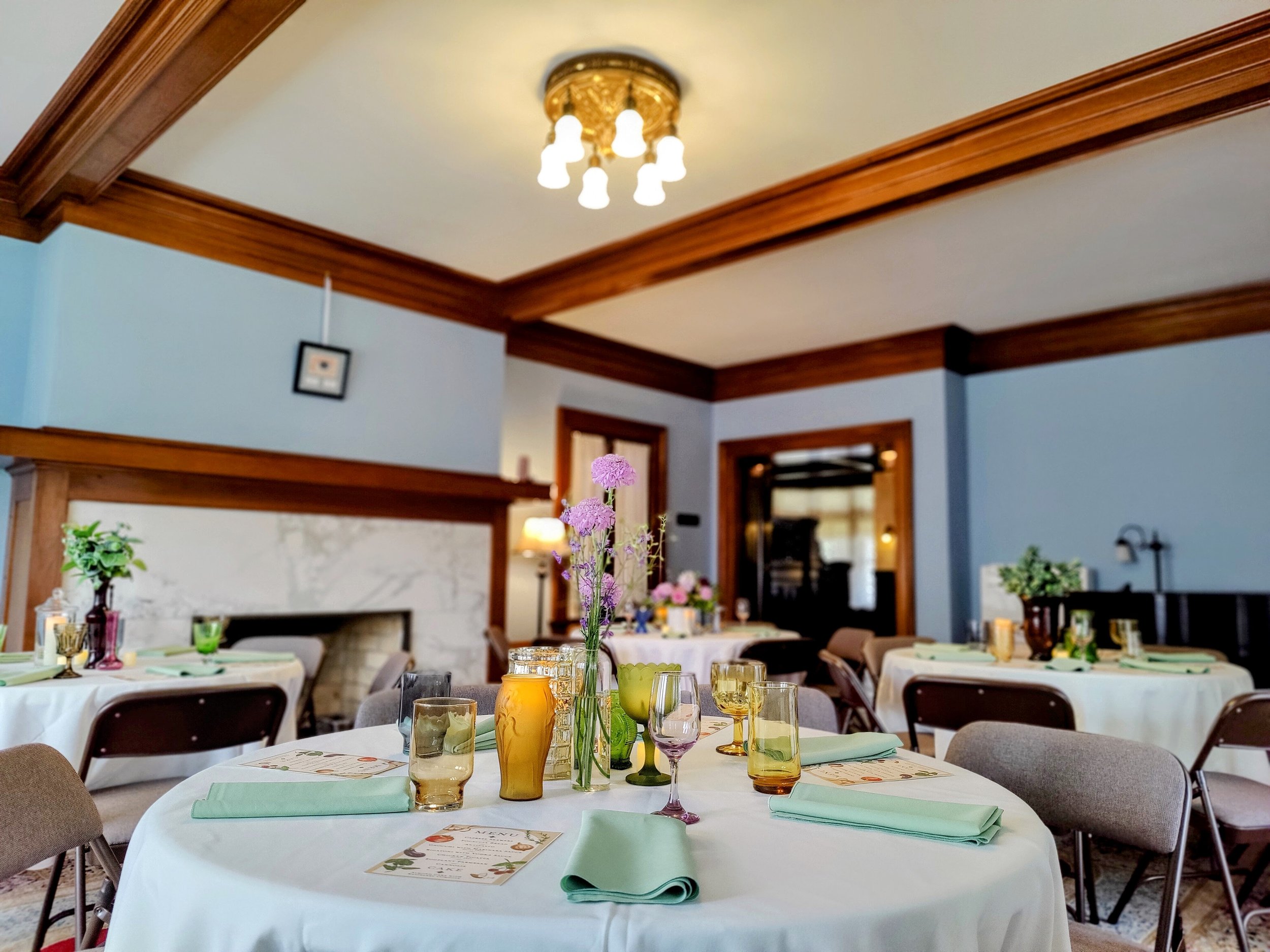 Elegant dining room with round tables set for an event, featuring white tablecloths, green napkins, and various colored glasses. Vases with flowers and decorative menus are on the tables. Walls are blue with wooden trim, and a chandelier overhead add