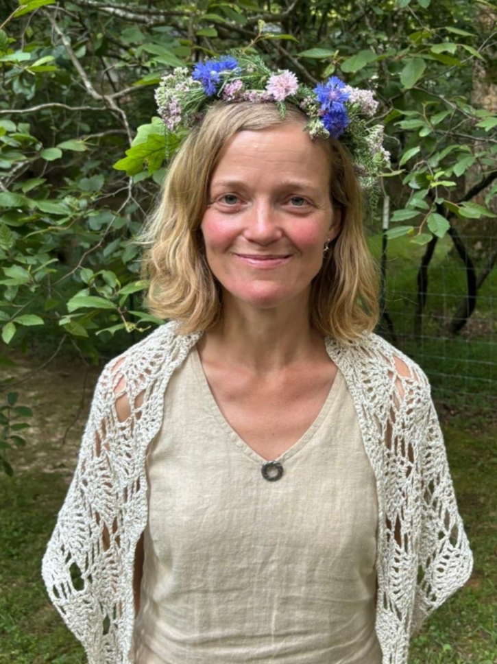 Woman with shoulder-length blonde hair wearing a flower crown made of blue, purple, and white flowers, and a beige crochet cardigan over a light-colored top, standing outdoors in a green, leafy area.