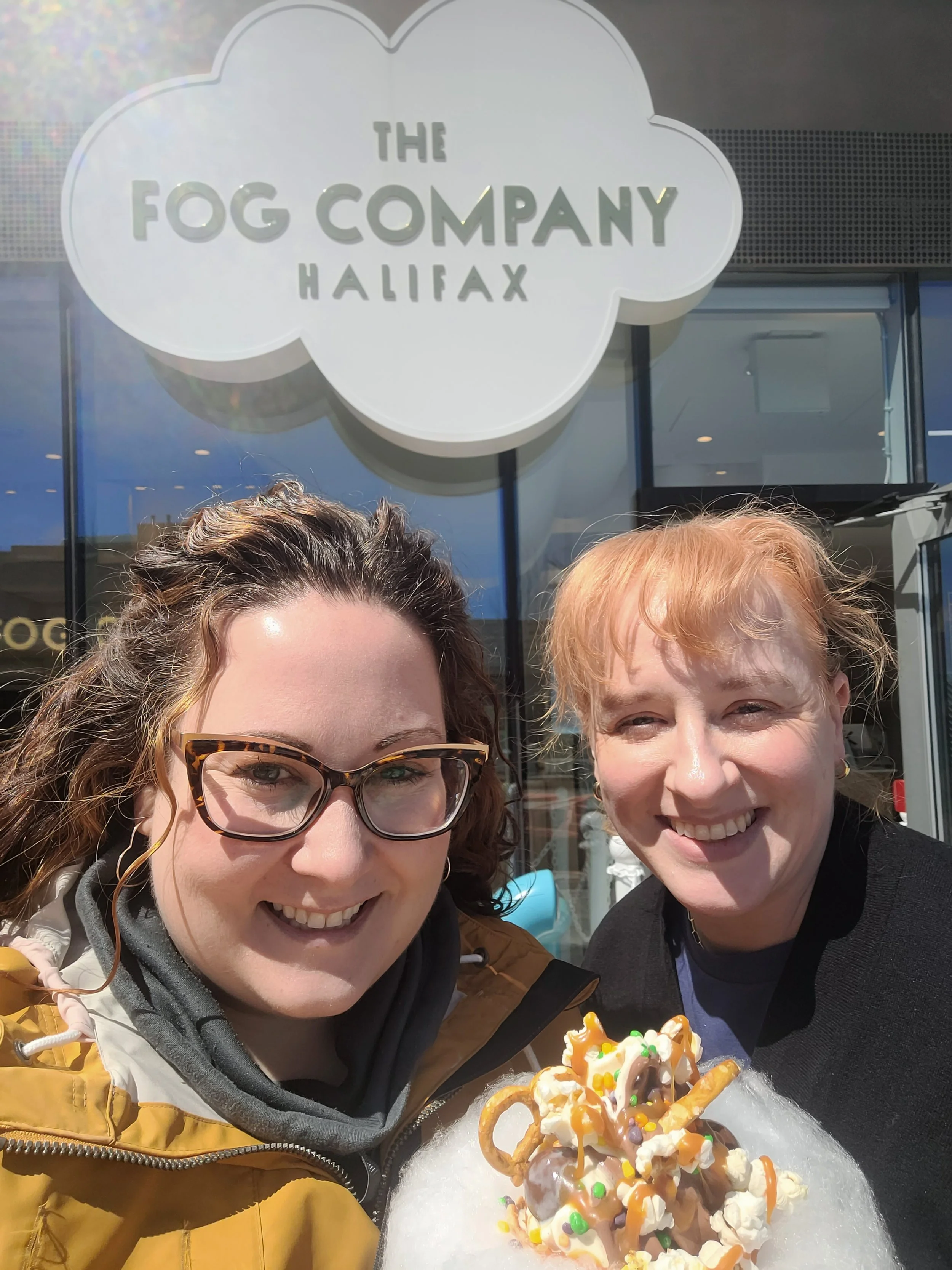 Two people smiling in front of The Fog Company Halifax sign, holding a dessert with pretzels, popcorn, and sprinkles on cotton candy.