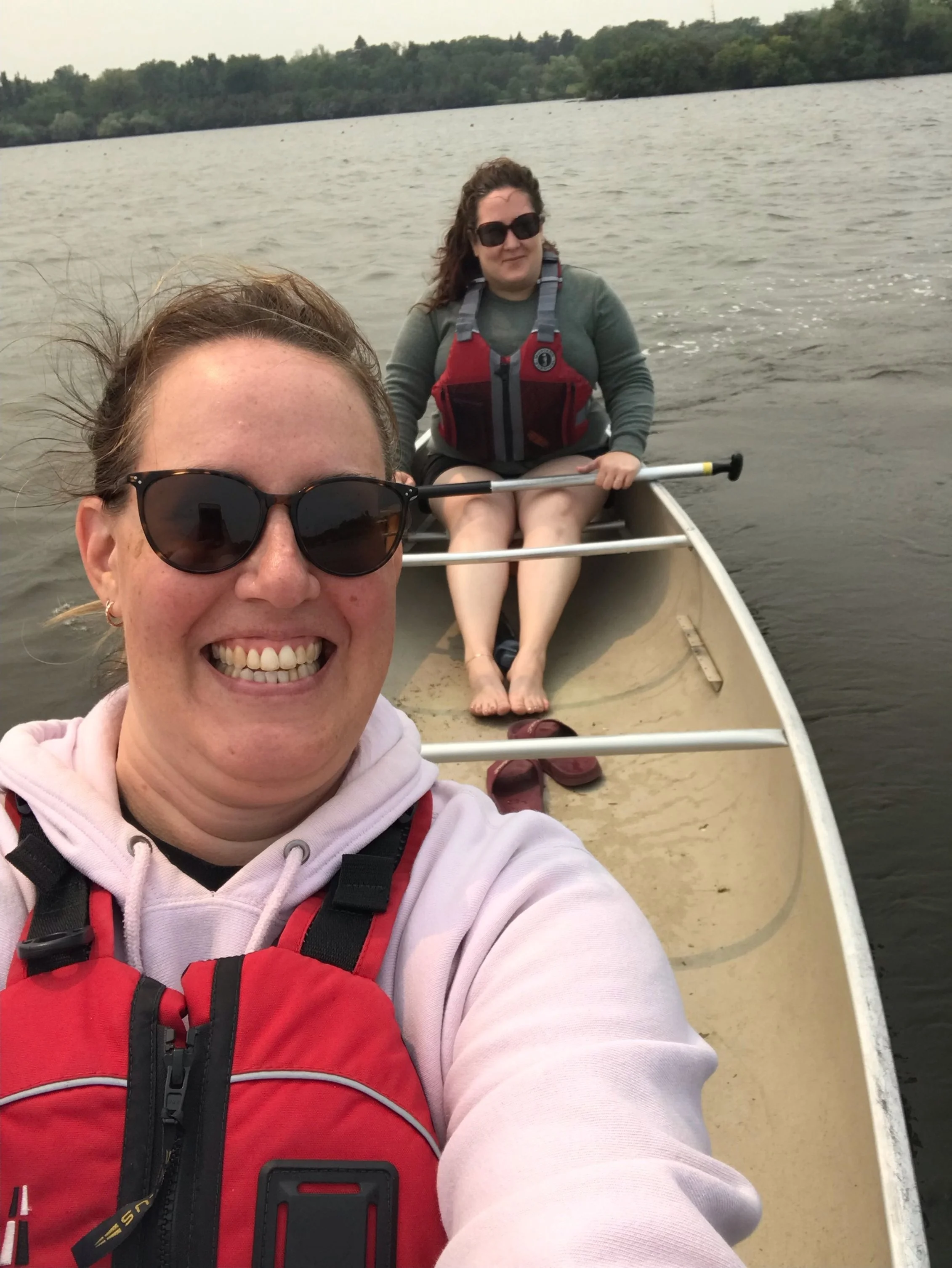 Two women smiling while canoeing on a lake, wearing red life vests, with trees in the background.