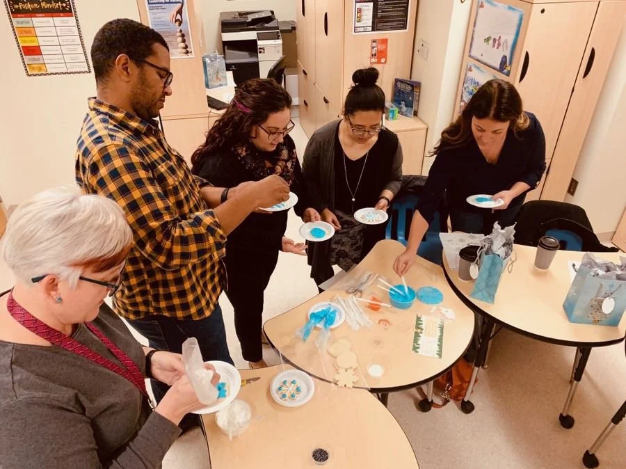 Group of people decorating cookies at a table in a classroom setting.