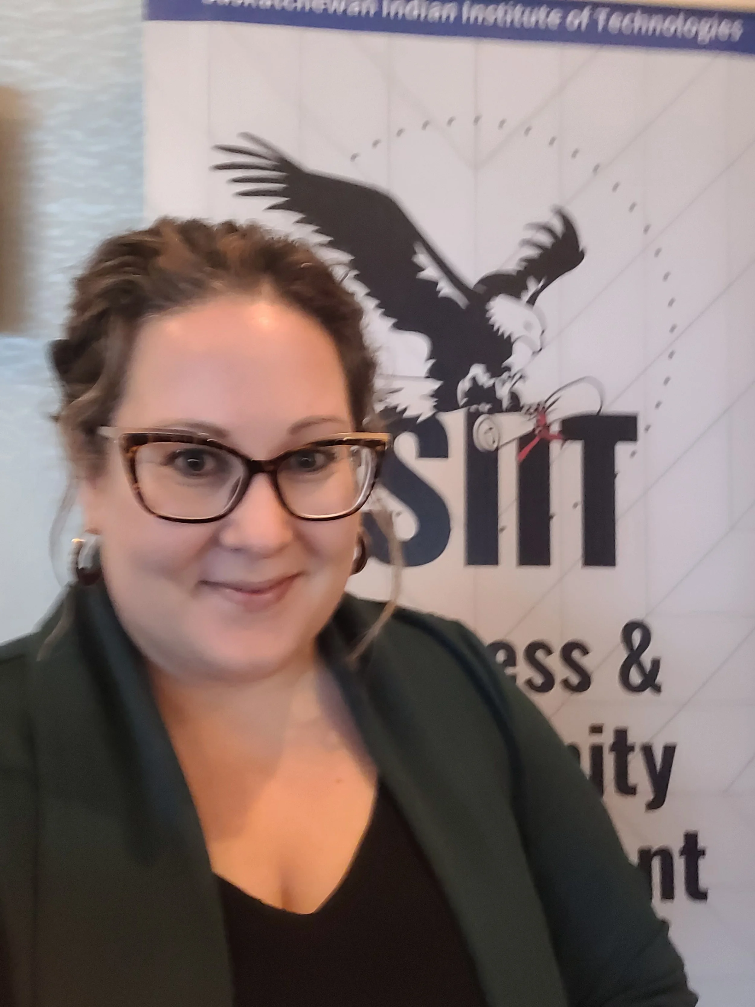 Person wearing glasses in front of a banner for the Saskatchewan Indian Institute of Technologies with an eagle logo.
