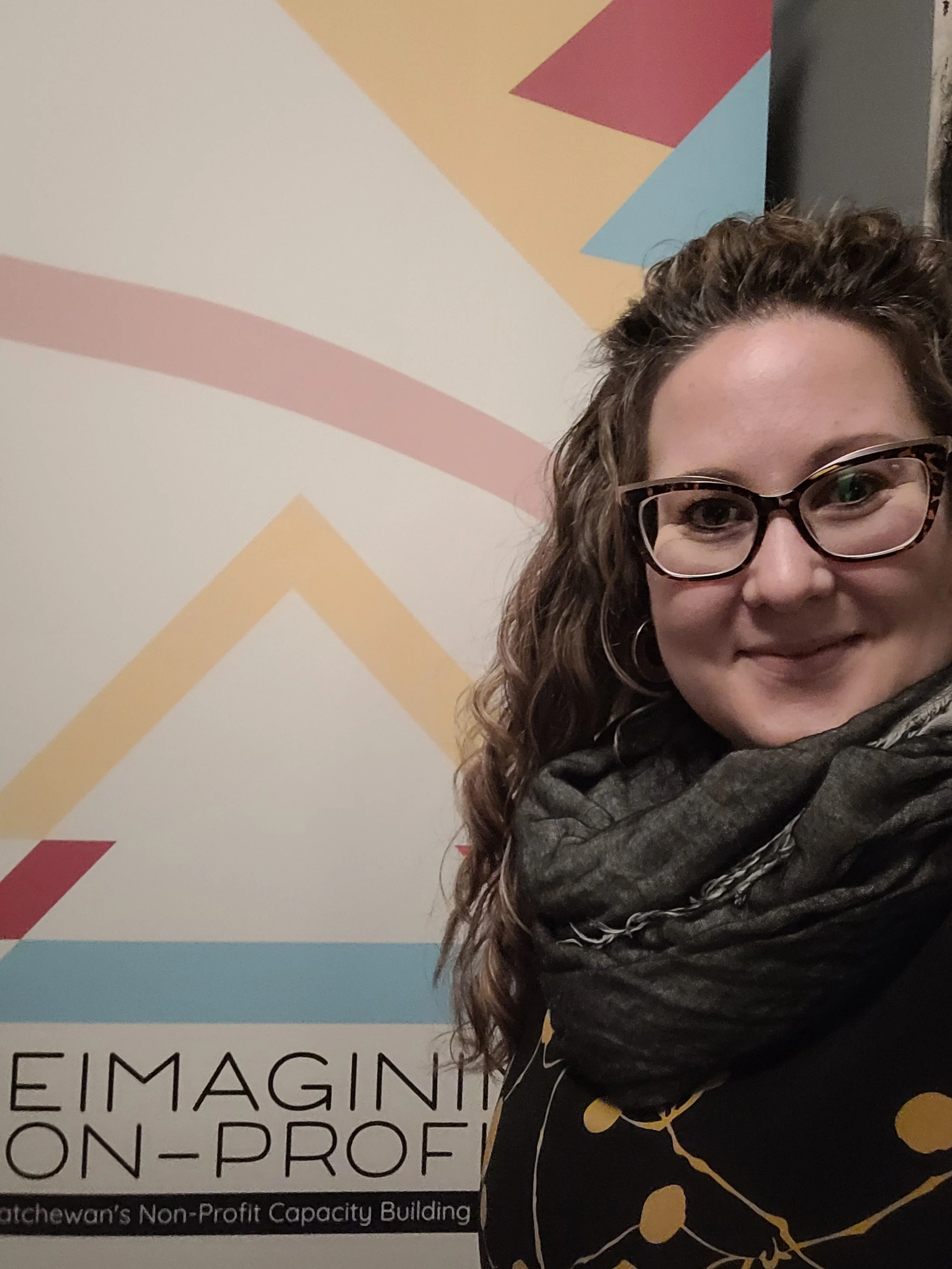Smiling person with glasses and curly hair standing in front of a sign with abstract design and text about nonprofit capacity building.