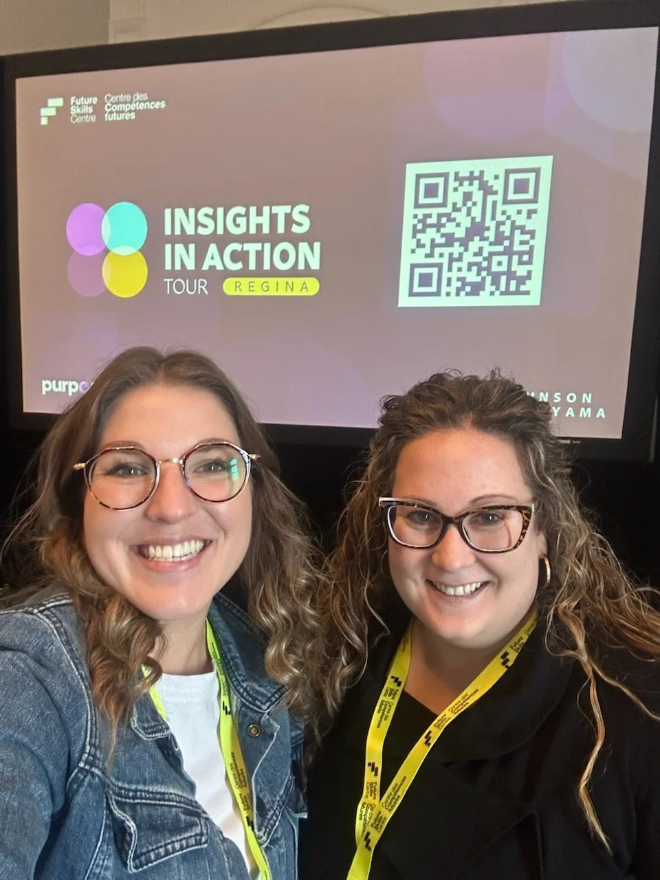Two women smiling in front of a screen displaying 'Insights in Action Tour Regina' with a QR code, wearing glasses and event lanyards.