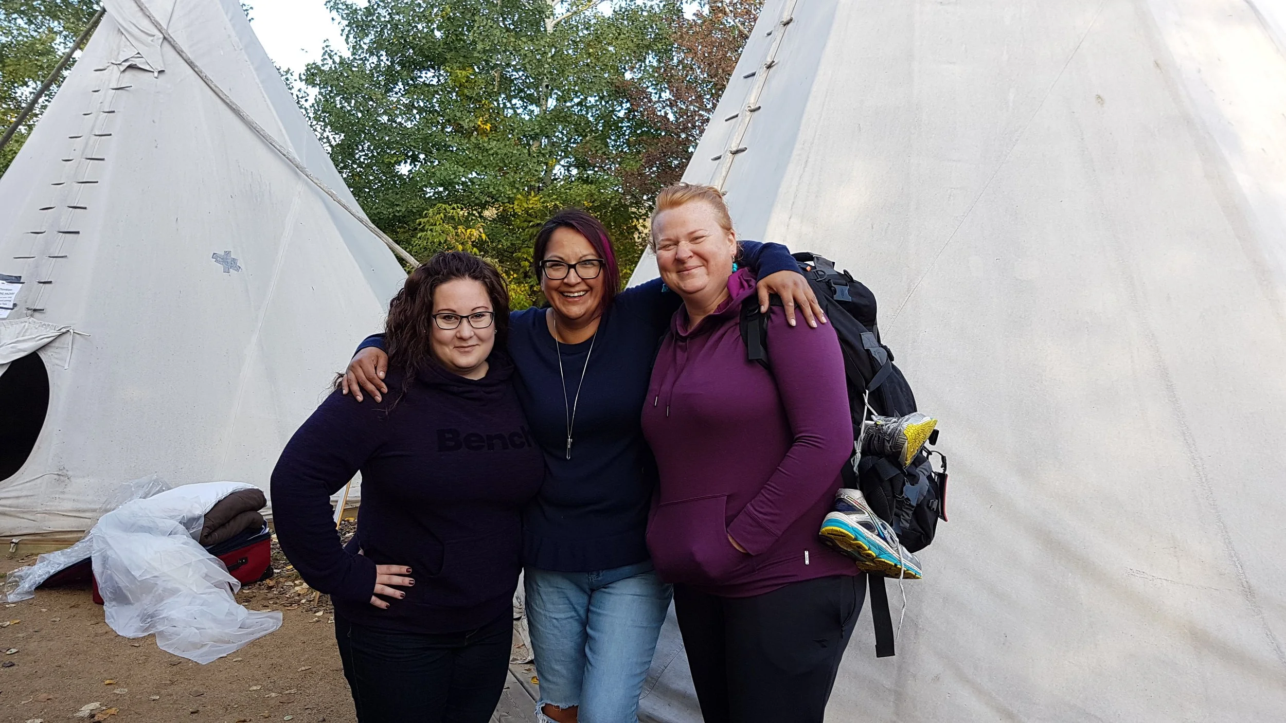 Three women smiling in front of large white teepees, surrounded by trees.