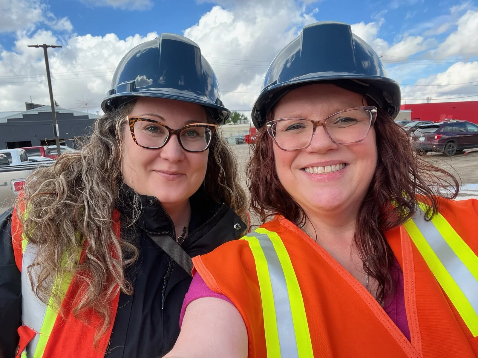 Two women wearing hard hats and orange safety vests smiling outdoors.