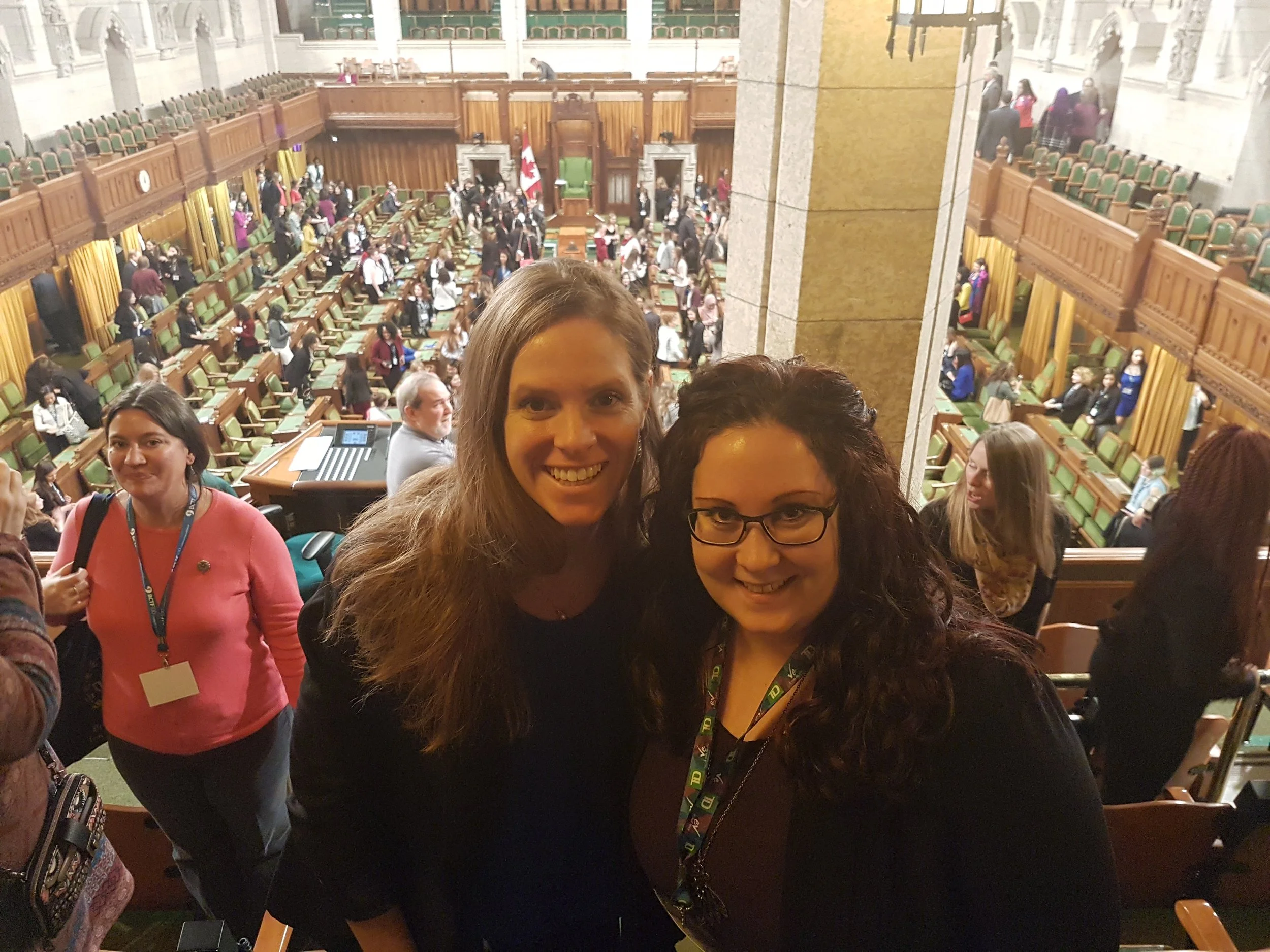 Two women smiling in the foreground in a large hall with people and rows of green seats in the background, possibly in a legislative chamber.