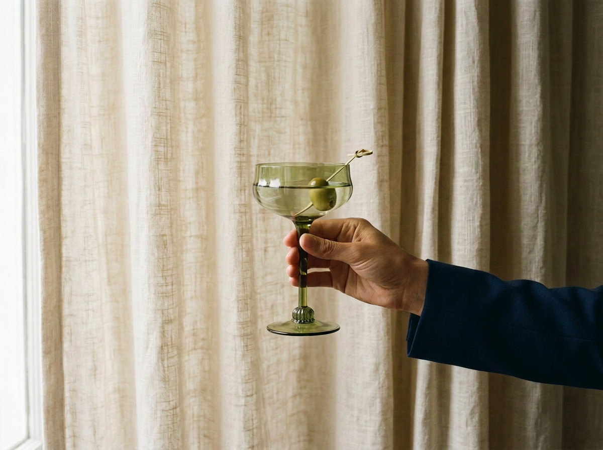 A hand holding a green martini glass with two green olives on a pick, against a beige curtain background.