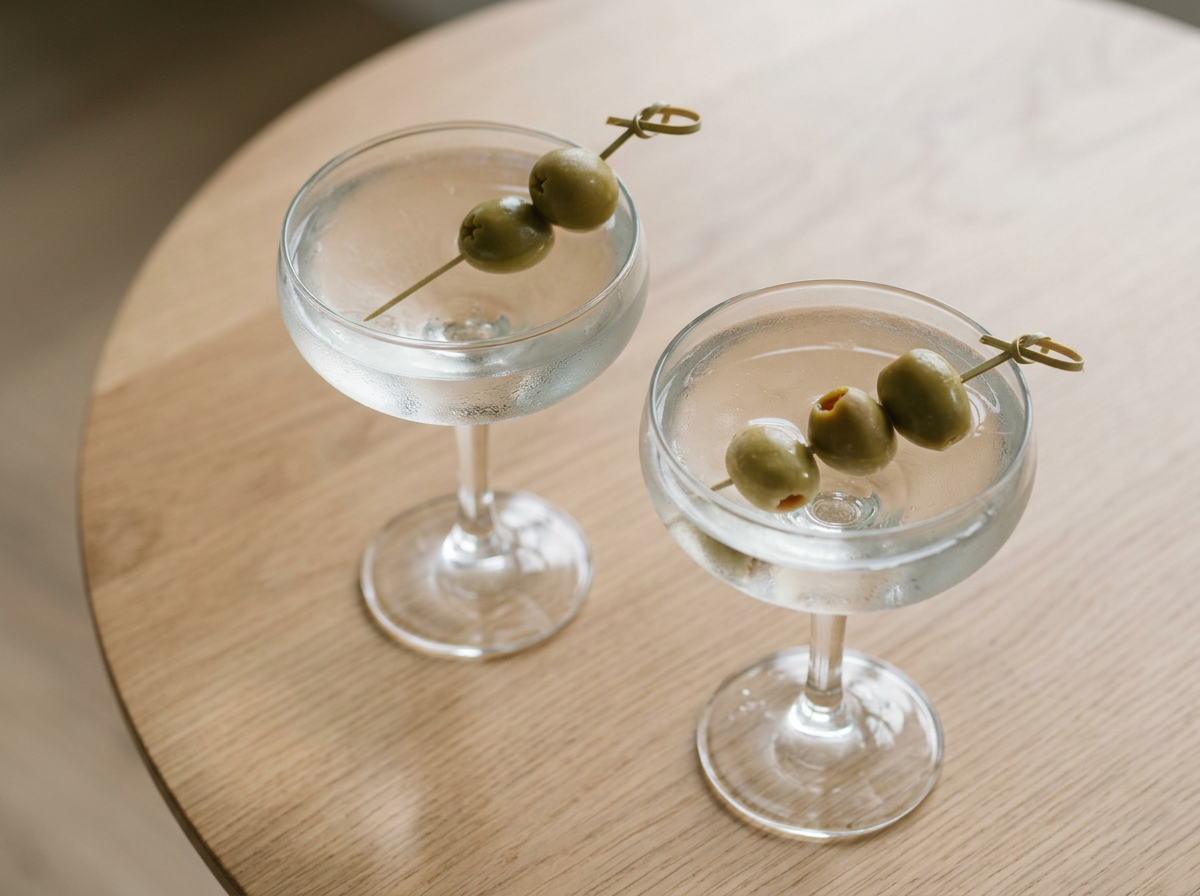 Two martini glasses filled with clear liquid and garnished with green olives on a skewer, placed on a light wooden surface.