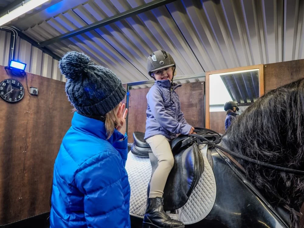 A child sitting on a black horse in an indoor riding arena, wearing a helmet and smiling, with an adult in a blue jacket nearby.