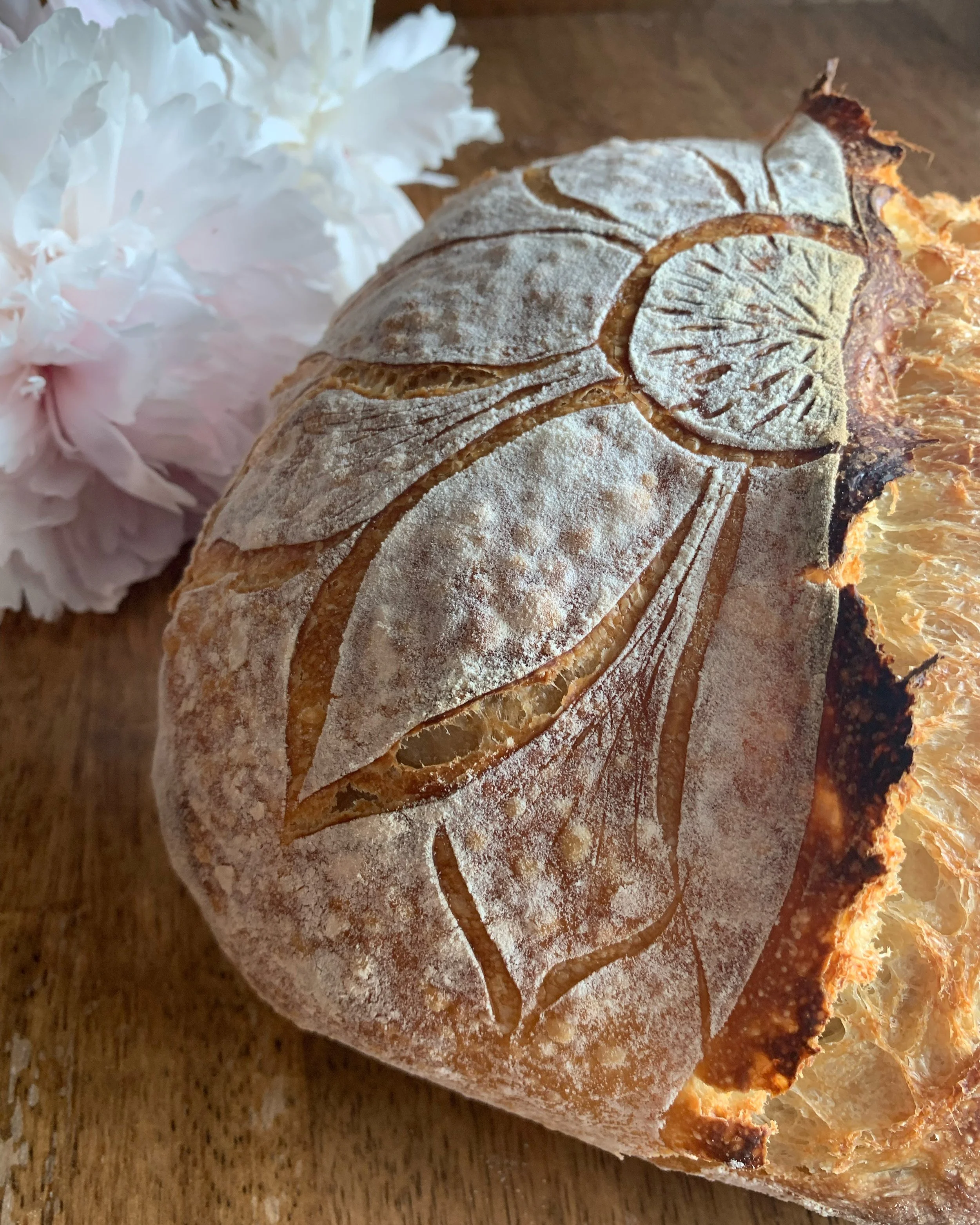 Close-up of a loaf of bread with a decorative leaf pattern on top, dusted with flour, next to pink and white peonies on a wooden surface.