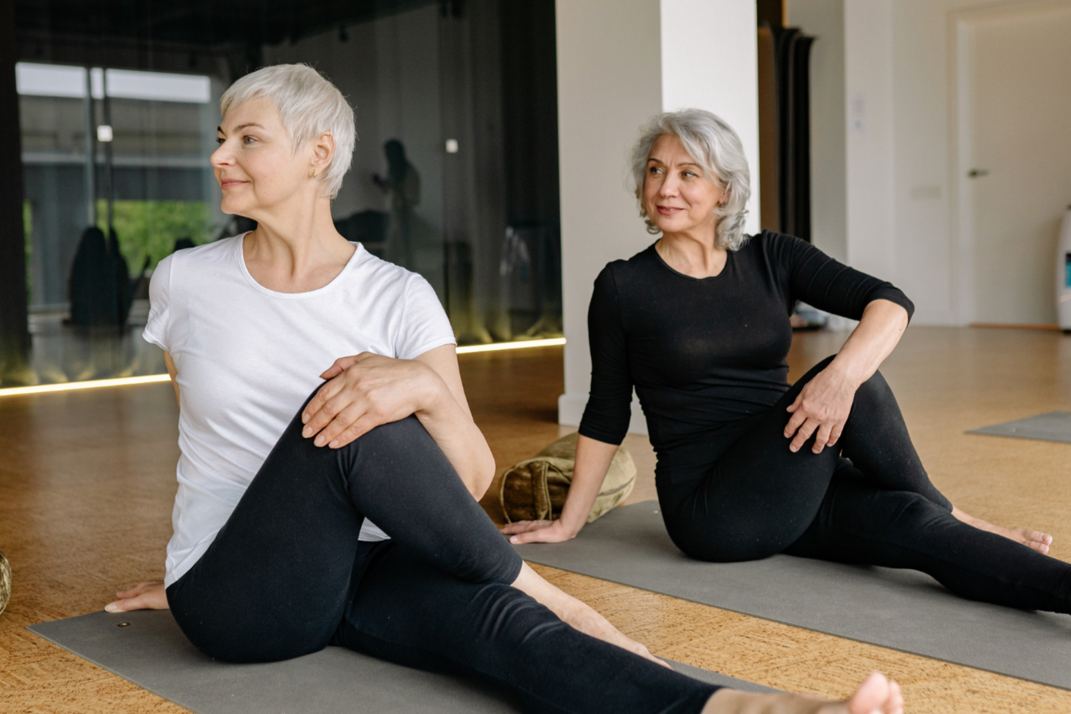 Two older women practicing yoga on mats in a wellness studio, sitting on their sides with one hand on their bent knees and the other supporting their back, both smiling and looking away.