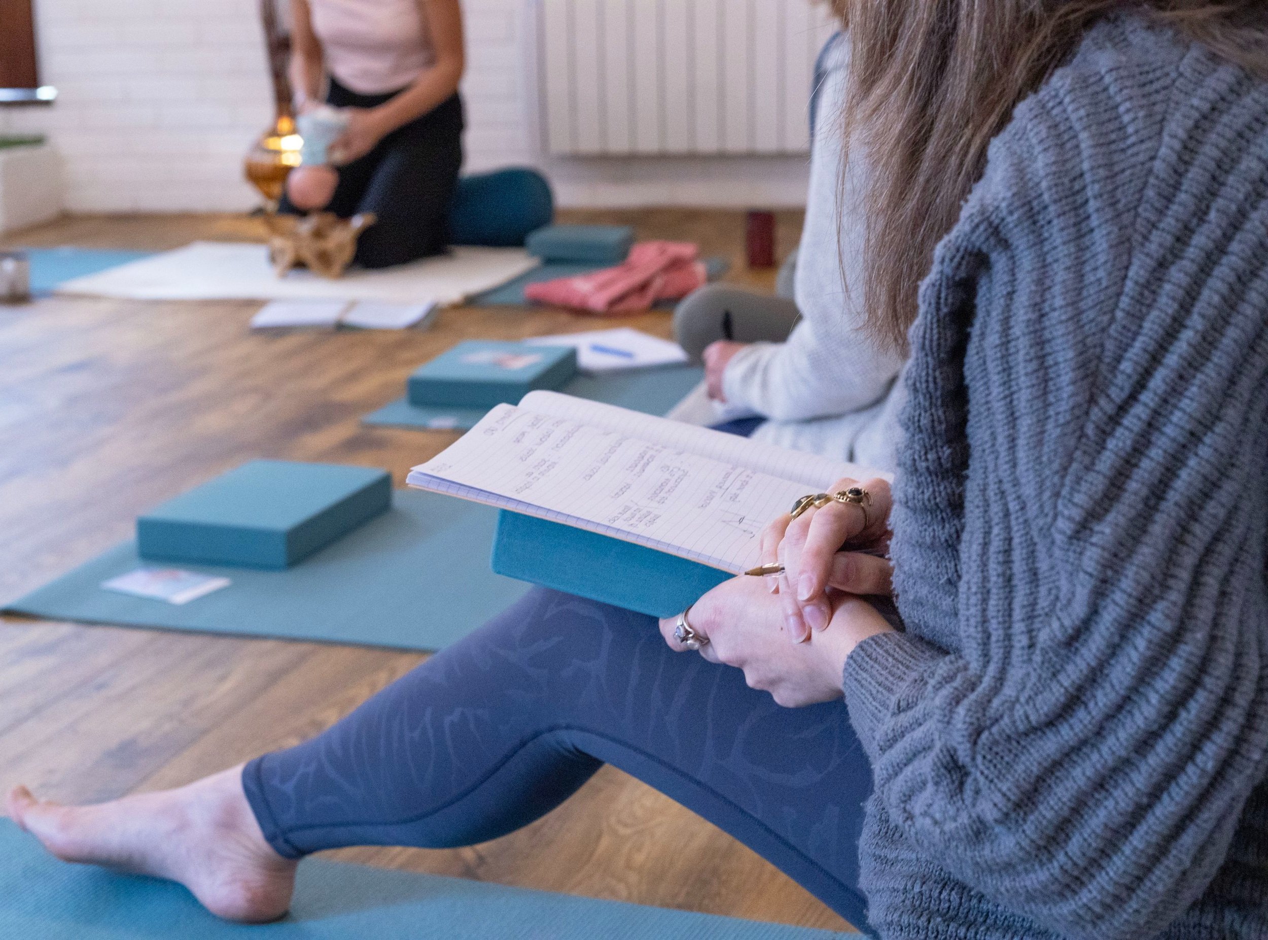 Close-up of a woman sitting cross-legged on a yoga mat, holding a notebook and pen, attending a yoga class with other participants and a dog in the background.